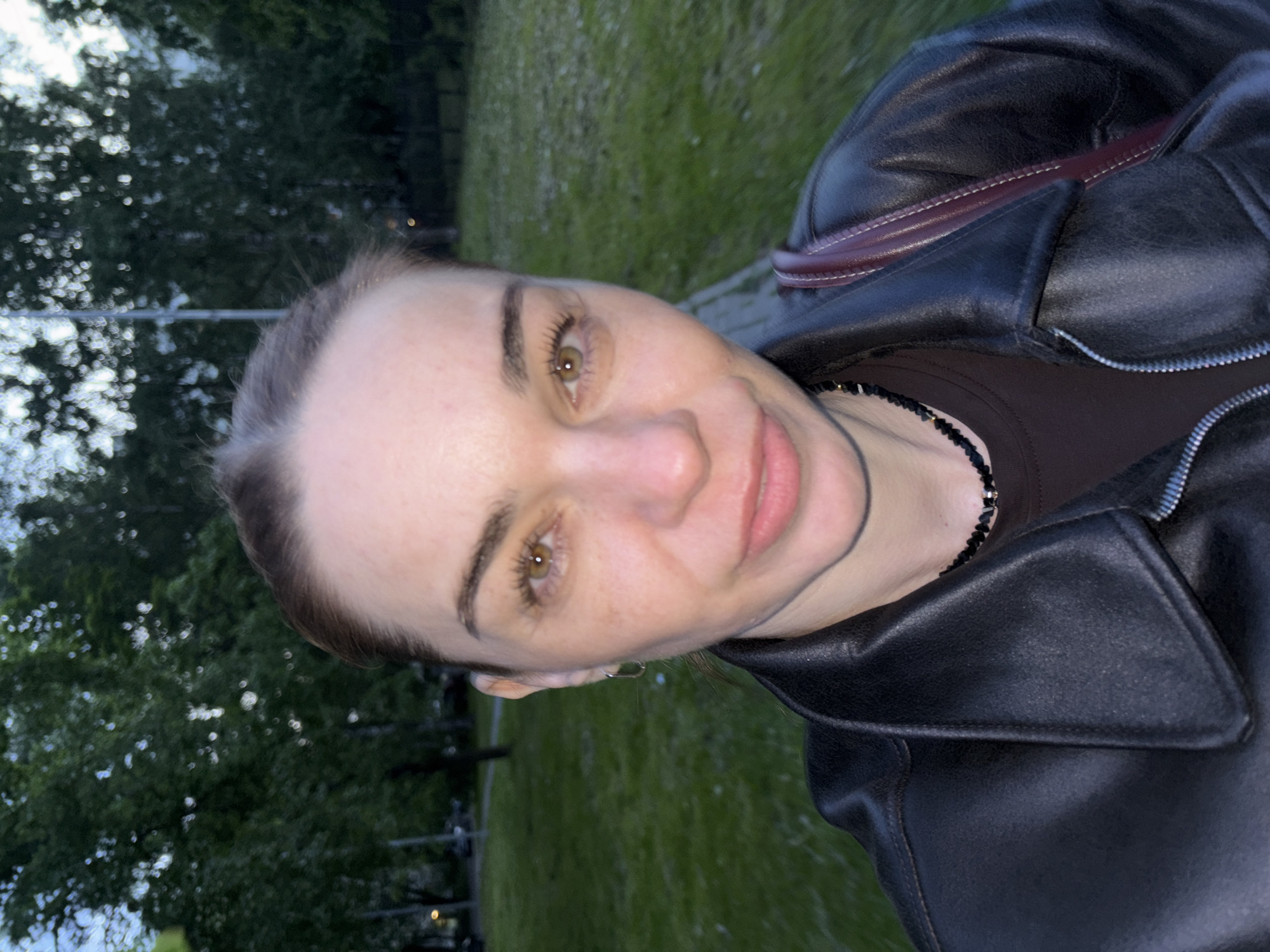 A close-up selfie captures a fair-skinned woman outdoors in Lednevo, Russia, likely during dusk or early evening due to the dim, natural light. The image is oriented sideways. She has light brown hair pulled back, prominent eyebrows, and striking hazel eyes that directly engage the viewer. Her lips are slightly parted in a soft, neutral expression or subtle smile. A few freckles are visible across her nose and cheeks.
She is wearing a dark, possibly black, leather jacket with a zippered front, over a dark brown top. A black beaded necklace is visible around her neck. A burgundy strap, likely from a bag or purse, runs across her right shoulder, contrasting with the dark jacket.
The background is softly blurred, showcasing green grass and dark, leafy trees, suggesting a park or natural outdoor setting. The bokeh effect in the background implies artificial lights or bright spots in the distance. There is no visible text in the image.
