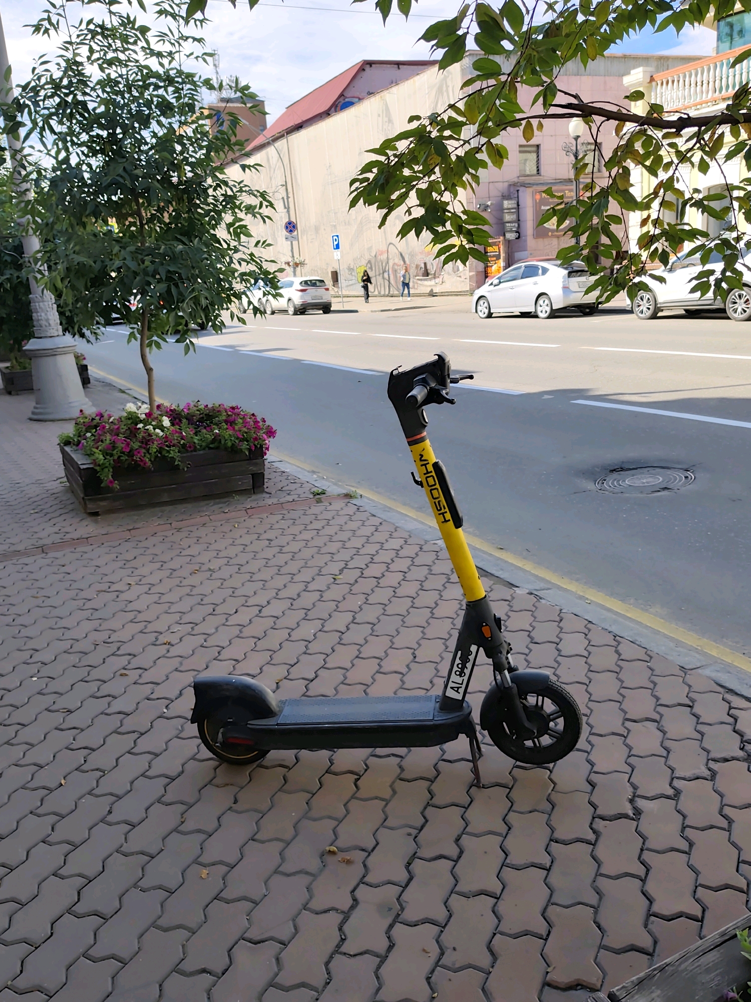 A yellow and black electric scooter is parked on a brick-paved sidewalk next to a street in Irkutsk, Russia. The scooter has "WHOOSH" written vertically on its yellow stem. The sidewalk is bordered by a street with a yellow line and white lane markings. A large planter box filled with pink and white flowers is located behind the scooter. In the background, there are buildings and trees. The sky is partly cloudy. Several cars are visible on the street, and a person is walking on the sidewalk in the distance. The overall scene appears to be a typical urban streetscape on a bright, sunny day.