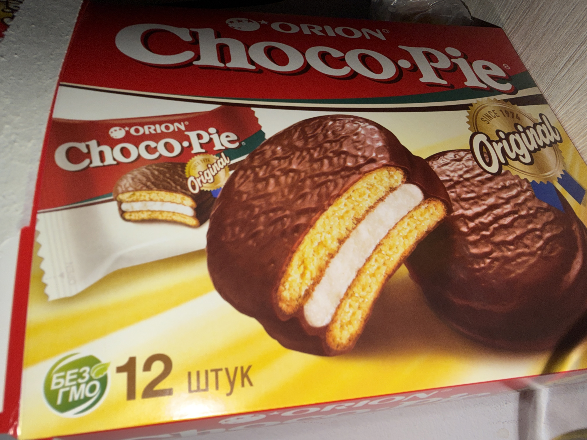 This is a close-up, eye-level shot of the front of a box of Orion Choco-Pie snacks. The box is predominantly red and yellow, with prominent branding. The main subject is the Orion Choco-Pie branding, featuring the "Choco-Pie" logo in large, white, stylized lettering against a red background. Below this, the text "Original" is displayed in a cursive font, encircled by a golden, scalloped design with the text "SINCE 1974" above it.
Two Choco-Pie snacks are visible on the packaging. One is shown whole, with a rich, dark chocolate coating and a visible creamy white filling between two golden cake layers. Another snack is depicted cut in half, revealing the layered interior.
In the lower left corner, there is a green circular logo with white text that reads "БЕЗ ГМО" which translates to "GMO free." To the right of this logo, the number "12" is displayed in large, bold font, followed by the word "штук," indicating 12 pieces. The background of the lower portion of the box is a bright yellow.
The overall impression is a colorful and appealing product advertisement, focusing on the dessert itself and its key attributes. The image appears to be taken indoors, possibly in a retail setting, given the lighting and the edge of a shelf visible at the top. There are no people or animals present in the image.