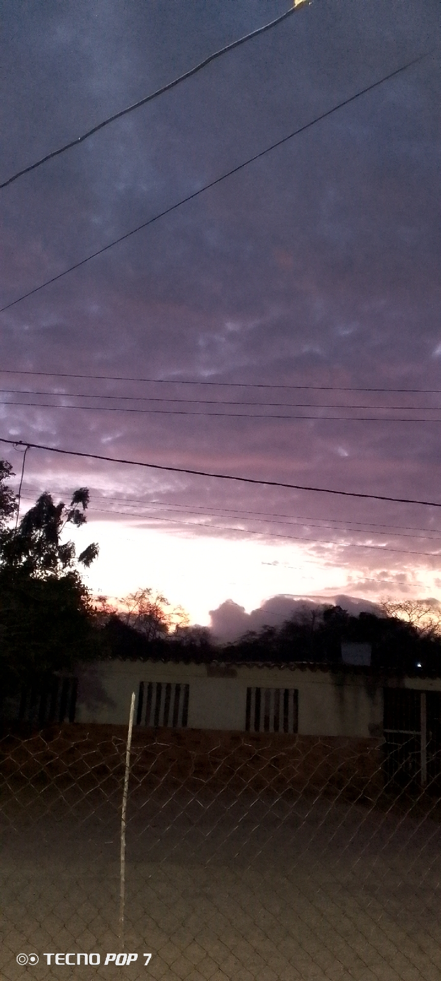 The image depicts a twilight scene in Altagracia, Venezuela. The sky is dominated by dark purple and grey clouds, with a band of brighter pinkish-orange light visible near the horizon. Several telephone wires are strung across the upper portion of the frame. Below the wires, silhouetted trees and a low, white building with barred windows are visible. A chain-link fence occupies the foreground. The bottom left corner of the image includes the watermark "TECNO POP 7".