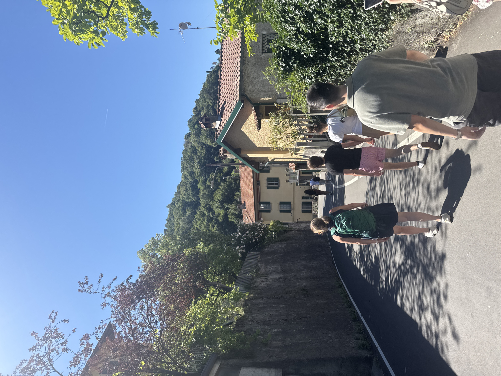 In Gittana, Italy, on a sunny day, a group of people are walking down a paved path lined with trees. The scene is dominated by a clear blue sky and lush green foliage. To the left, a hillside covered in dense trees rises sharply, with houses nestled among the greenery. Some of the houses have terracotta tiled roofs. To the right, the path is shaded by the dappled sunlight filtering through the leaves of trees.
The people are casually dressed, suggesting a leisurely activity. One person is wearing a green backpack and dark shorts. Another is in a dark t-shirt and pink and white striped shorts. Further up the path, other individuals are visible. The overall atmosphere appears relaxed and pleasant, indicative of a warm, clear day. The sun is high in the sky, casting long shadows, suggesting it is likely midday or early afternoon. There is no visible text in the image.