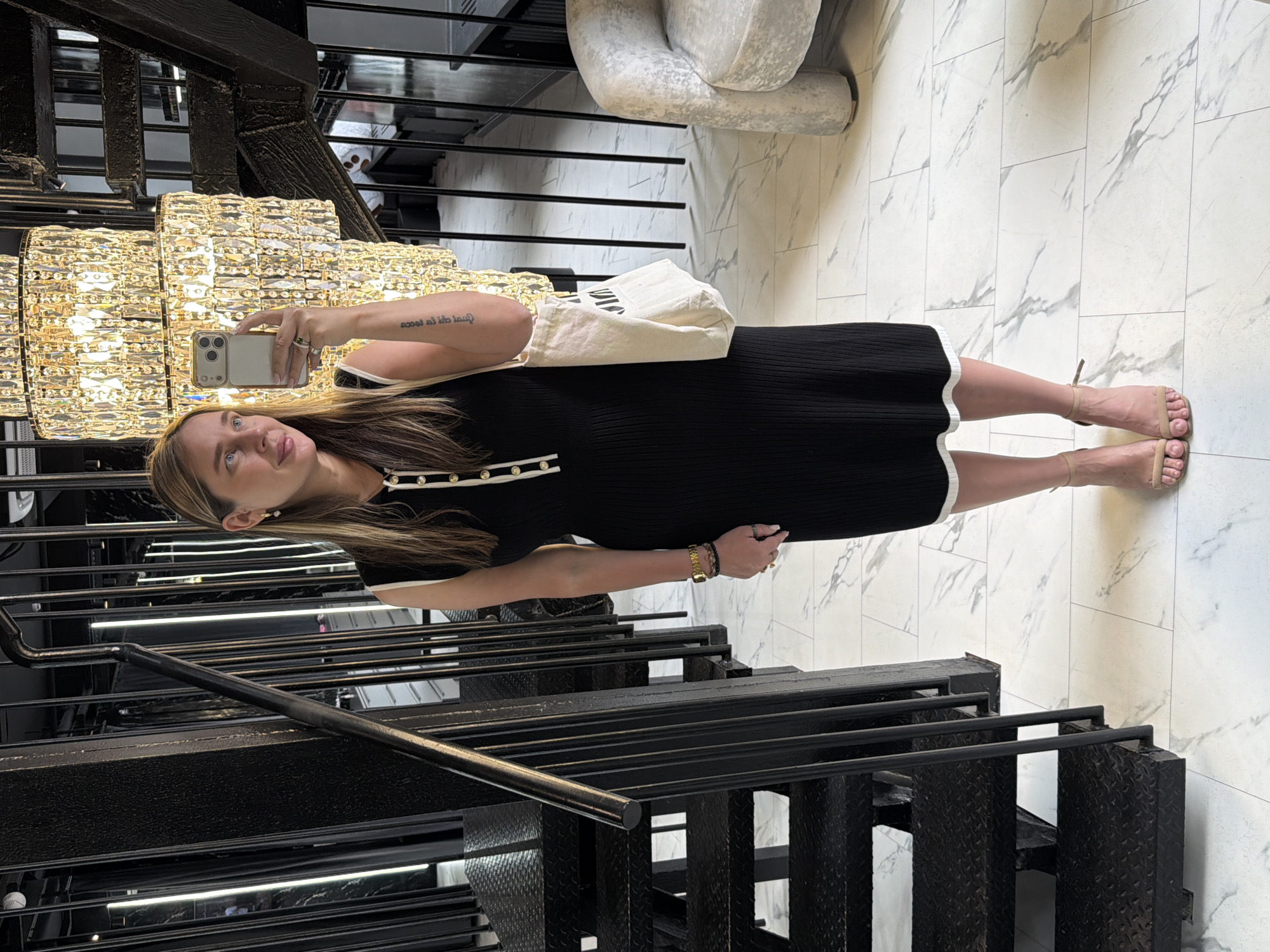 A woman is standing on a staircase in what appears to be an indoor location, possibly a retail space or residential building. The scene is illuminated by a large crystal chandelier. She is holding a smartphone, likely taking a selfie. She is wearing a black dress with white trim, a watch and bracelets on her left wrist, and sandals. She has shoulder-length hair and is looking at the phone. She is carrying a white bag over her shoulder. The environment features modern architectural elements, including black metal stairs and railing. The flooring appears to be white marble. There is a sculpture-like chair in the background.
There is insufficient information to establish with certainty that this is Moscow, Russia. The provided image lacks specific architectural landmarks or signage that definitively place it in Moscow.