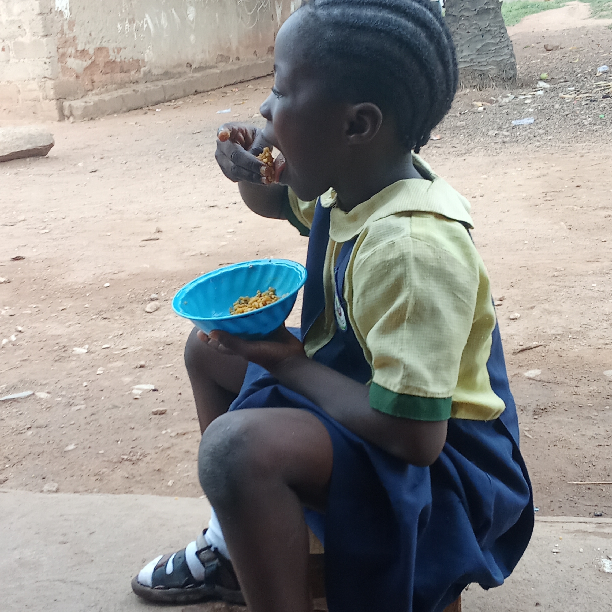 Akwanga, Nigeria: A young girl in a school uniform, seated outdoors, eats from a blue bowl with her hand. The uniform is two-toned, with a yellow upper section and a dark blue lower section. The girl wears sandals and white socks. The background includes a building and dirt ground.