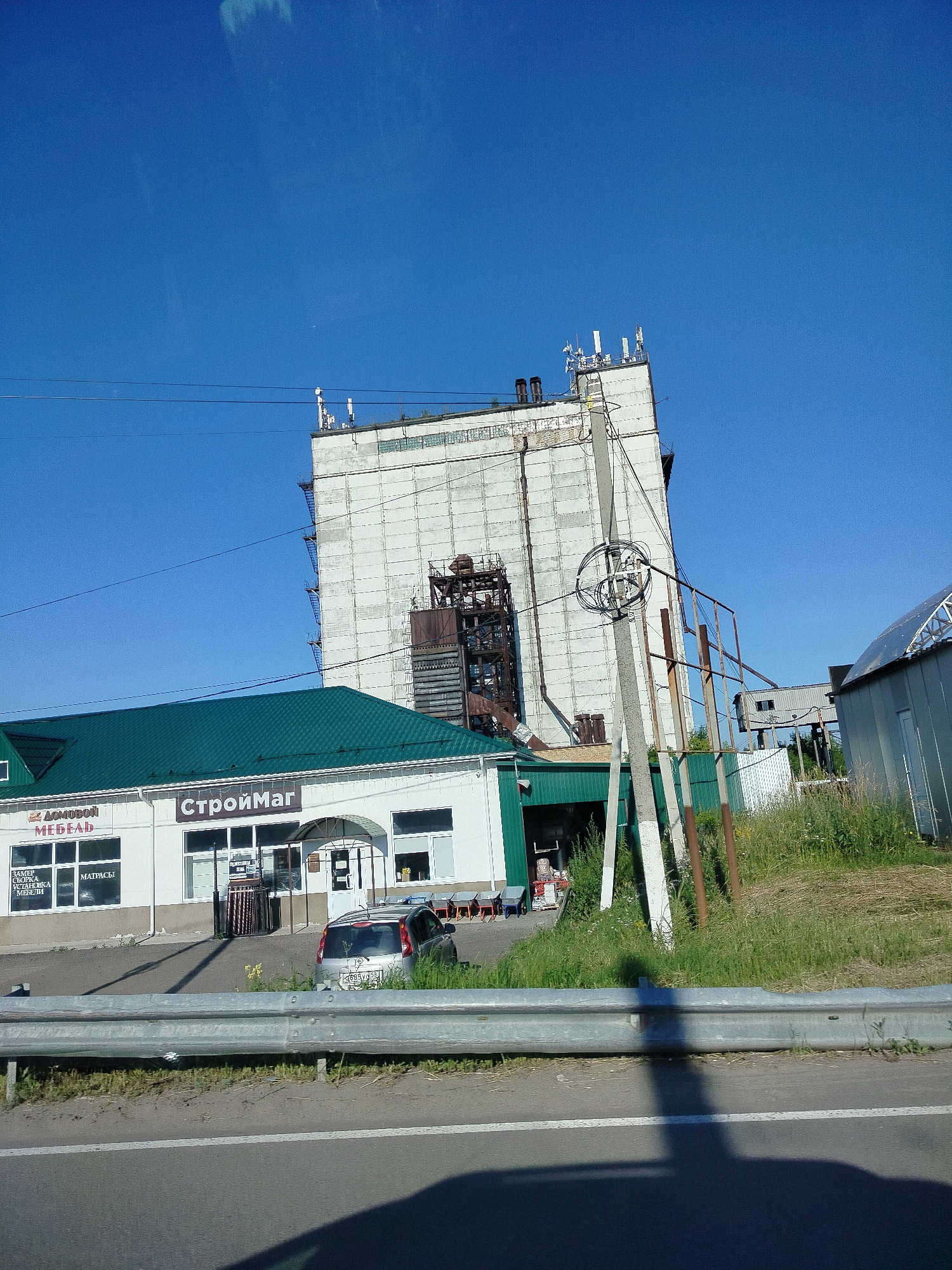 The image shows a sunny day in Grabovo, Russia. The main subjects are buildings and a silver car parked in front of a store. The scene is set along a road, indicated by the guardrail in the foreground and what appears to be a highway line.
The buildings consist of a large, light-colored industrial structure with visible scaffolding or external staircases and a smaller building in front of it with a green, sloped roof. The smaller building appears to be a retail store, as suggested by the visible signage and windows displaying merchandise or services. The industrial building has several antennas on its roof, indicating it might be a telecommunications tower or part of a larger industrial complex.
The text visible on the smaller building includes "СтройМаг" and "ДОМОВОЙ МЕБЕЛЬ". There are also smaller texts on the windows, possibly related to services offered, such as "ЗАМЕР СБОРКА УСТАНОВКА МЕБЕЛЬ" and "МАТРАСЫ".
The time of day is likely afternoon, given the bright sunlight and the presence of shadows. The weather is clear and sunny, with a bright blue sky. The overall atmosphere is mundane and functional, depicting a typical commercial and industrial area. There are no people visible in the image, and the car appears to be stationary. The grassy area and the vegetation suggest a setting outside of a densely populated urban center.