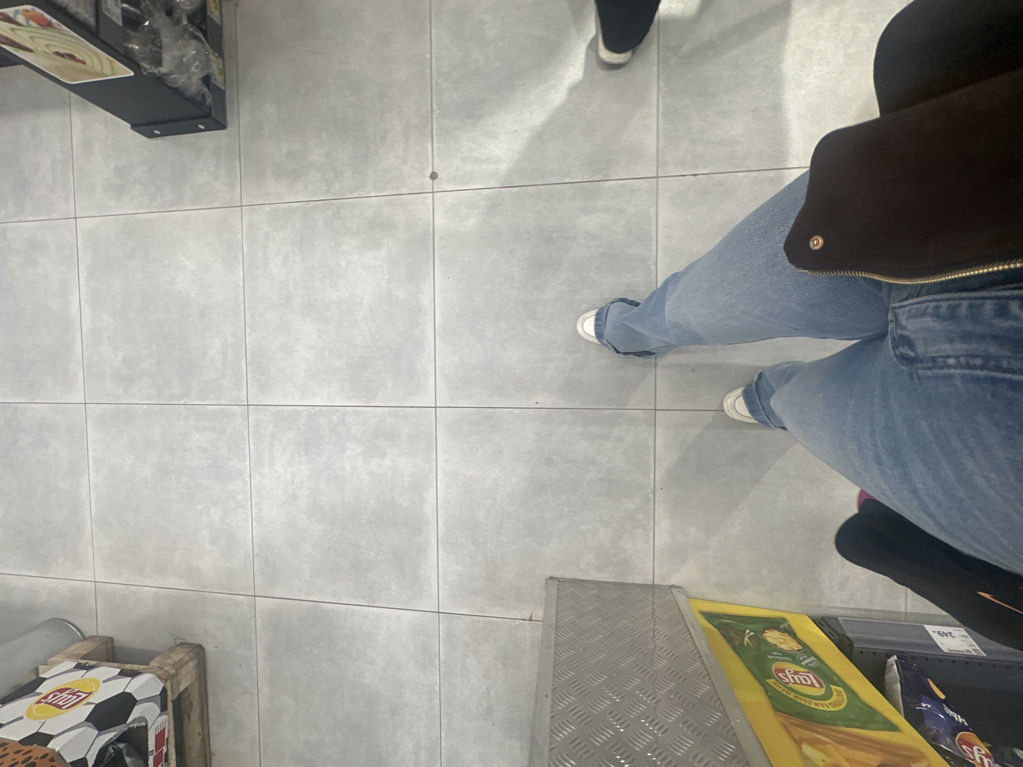 The image shows a person standing in what appears to be a retail store aisle, possibly in the city of Megion, Russia. The person is wearing light blue jeans and white sneakers. Part of a dark-colored jacket is also visible. Nearby are displays containing snack food products, including Lays potato chips. The floor is tiled in a light gray color. Another person is also present to the right of the view in the image.
