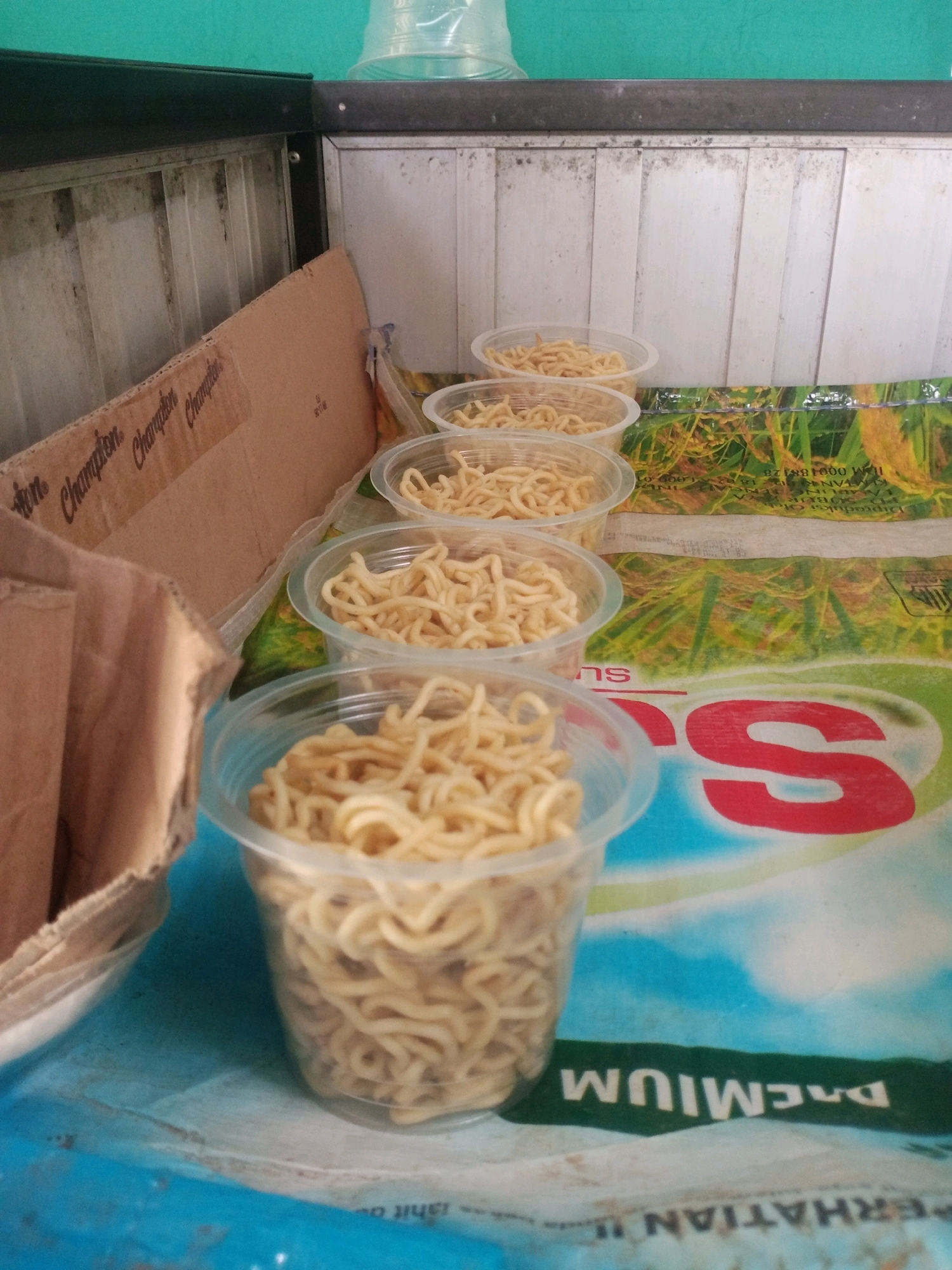 The image shows a close-up view of several plastic cups filled with instant noodles. The cups are arranged in a line, with the closest cup prominently featured in the foreground. The noodles appear to be dry and uncooked. In the background, there is a cardboard box with text that reads "Champion". The surface beneath the cups and box is a patterned material, possibly a tablecloth or mat, with green, yellow, and blue elements. There is a green wall in the upper background. The lighting suggests an indoor setting. The image does not provide any specific information about the location in Bandar Lampung, Indonesia, beyond the possibility that it could be a food stall or a home kitchen.