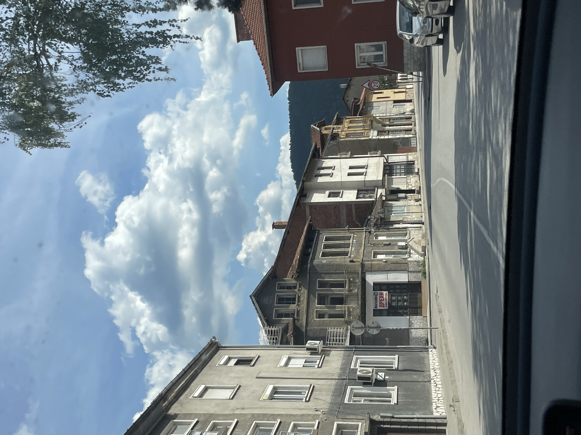 This is a street view captured from inside a vehicle, looking out at a row of buildings on the left side of the road. The buildings are of varying architectural styles and ages, with some appearing to be older, two- and three-story structures, and others more modern. The facades are primarily light-colored, with some featuring gray plaster, white trim around windows, and dark-colored doors. One building on the left has multiple air conditioning units mounted on its exterior. A sign with Cyrillic script and an image of a store is visible above a storefront on the left.
The street itself is paved and appears to be a two-lane road. A white line marks the center of the road, and a white curved line suggests a turning lane or a change in the road's direction. Sidewalks are visible along the buildings. Shadows from trees are cast across the road and sidewalk.
In the background, a dense line of green trees forms a natural landscape, with hills rising behind them. The sky is blue with scattered white, fluffy clouds. A speed limit sign indicating "20" is visible on a pole on the right side of the street, along with a triangular warning sign. Two cars, a silver sedan and a darker vehicle, are parked on the right side of the road. A portion of a tree with hanging leaves is visible in the upper right corner of the frame, partially obscuring the sky. The bottom of the image shows the dashboard or interior of the vehicle from which the photo was taken.