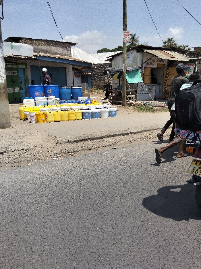 The image displays a daytime street scene in Mkomani ward, Kenya, characterized by roadside commerce and pedestrian activity. In the foreground, an asphalt road surface occupies the lower half of the frame, bordered by an unpaved dirt and gravel shoulder on the right.
Approximately two dozen large blue plastic barrels are stacked on the dirt shoulder to the left, with over one hundred smaller plastic containers, predominantly yellow, white, and light blue jerrycans and buckets, arranged in rows in front of them. A male individual wearing a white kufi and a dark shirt stands near the blue barrels, facing away from the camera.
In the midground, two individuals walk from right to left along the road shoulder. The closer person wears a dark backpack, dark shirt, and shorts, with visible bare feet or sandals, and is partially obscured by the rear of a motorcycle or scooter, identifiable by a partially visible Kenyan license plate "KM 869" on its fender. The second individual, slightly behind, wears a dark shirt and pants.
Several low-rise buildings line the street. The structure on the far left displays partial signage for "SafaCom Services" and "M-PESA." An adjacent building to its right features corrugated metal roofing and faded paint, with prominent signs reading "GENIUS PHONE REPAIR" and "FUNDS." A green tarp is affixed to its front. Utility poles with overhead electrical wires are visible on both sides of the street. The sky above is light blue with scattered white clouds.