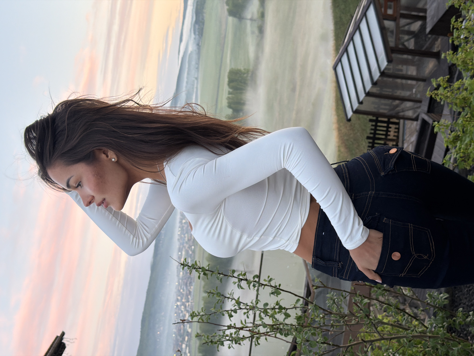 A woman with long brown hair, wearing a white long-sleeved top and dark blue jeans with a copper button detail on the back pocket, stands outdoors. Her left hand is in her pocket, and she is looking down and to her left. The background consists of a landscape with trees, fields, and what appears to be fog or mist in the distance. A building or structure with a glass roof is visible on the right side of the frame. The sky has hues of pink and orange, suggesting it is either dawn or dusk. There is foliage in the lower right and lower left corner of the image, partially obscuring the view. The city name of Shamoni, Russia is not discernible from the image.