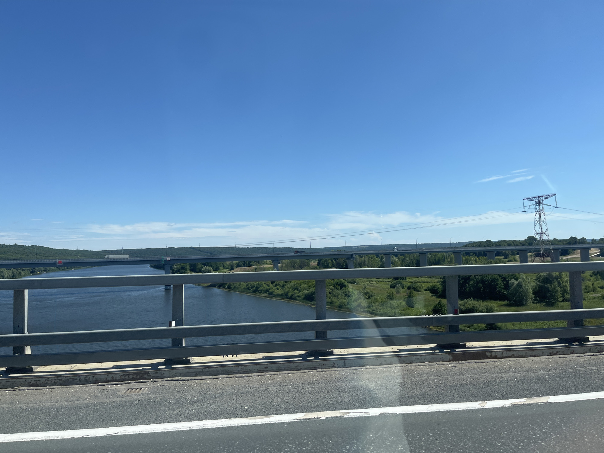 This media file is a landscape photo taken from a bridge, likely on a sunny day with a clear blue sky. The foreground features a gray metal railing and a section of asphalt road with a white line. Beyond the railing, a wide, dark blue river flows, bordered by lush green trees and vegetation on both banks. In the distance, multiple bridges span the river. The closest bridge in the mid-ground has several vehicles, including a white truck, visible on its deck. Further in the background, another bridge is visible, with a single white truck on it. A tall, lattice-style power line tower stands to the right of the image, with power lines stretching across the horizon. The overall scene suggests a rural or semi-rural location with significant infrastructure, possibly in Zasur'e, Russia. There is no visible text in the image. The time of day appears to be daylight, given the bright sky and strong shadows. The weather seems to be fair and warm.