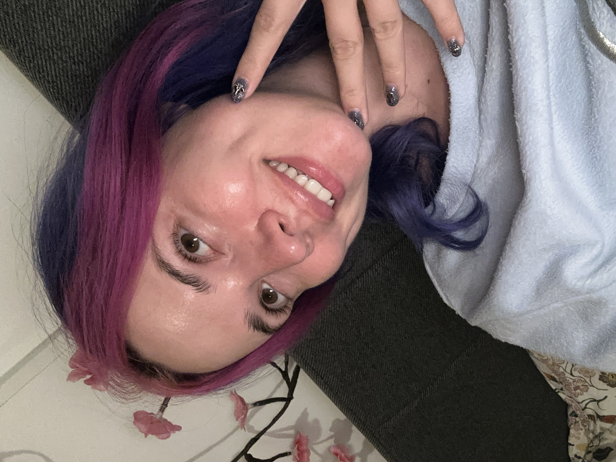 The media file presents a close-up, tilted portrait of a person, likely a woman, lying down and smiling broadly at the camera. The individual features striking two-toned hair, with vibrant purple on one side and bright magenta or pink streaks on the other. A hand is gently raised, partially obscuring the forehead, showcasing manicured nails painted with a dark grey, glittery design.
The person's expression is joyful and relaxed, with a wide smile revealing white teeth. They are wearing a light blue, soft-textured garment, resembling a towel or bathrobe, draped around their upper body. The head rests on a dark gray, upholstered surface, indicative of a couch or bed. In the lower left corner of the frame, a lighter surface is visible, adorned with what appears to be decorative pink flowers on a branch. The setting is indoors. While context indicates the City of Zagreb, Croatia, no specific visual cues confirm this location within the image. No text is visible.