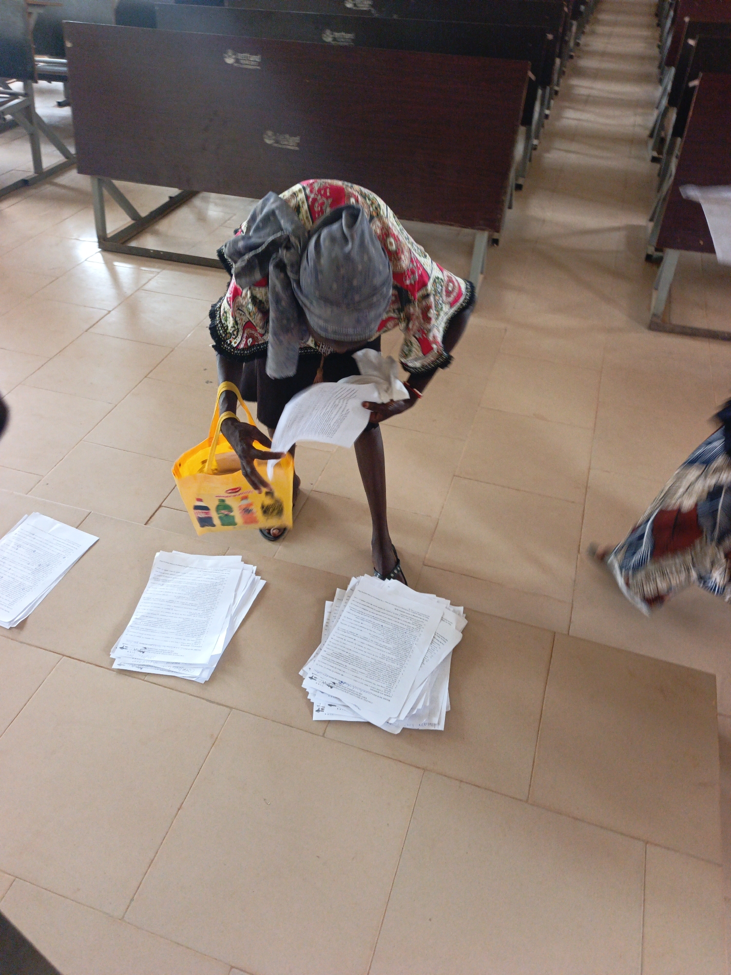 Akwanga, Nigeria. An individual stands in an indoor space, possibly a classroom or hall, surrounded by rows of dark-stained desks. The individual, dressed in colorful patterned clothing, holds papers in their hands and appears to be sorting or organizing them. A yellow shopping bag sits nearby, and stacks of papers are scattered on the tiled floor. The light is bright and appears to be natural.