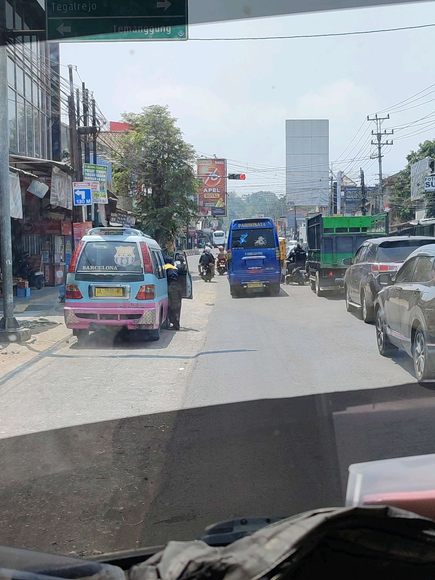 This is a street-level photograph taken from inside a vehicle in Secang, Indonesia. The scene depicts a moderately busy street with several vehicles, including a light blue and pink van with "BARCELONA" written on the back, a blue van labeled "PARIWISATA," a green truck, and several cars. A person in dark clothing stands beside the "BARCELONA" van with the door open. There are street signs indicating directions to "Tegalrejo" and "Temanggung." Commercial buildings and trees line the street, and overhead power lines are visible. The sky is bright and appears overcast. The lighting suggests it is daytime.