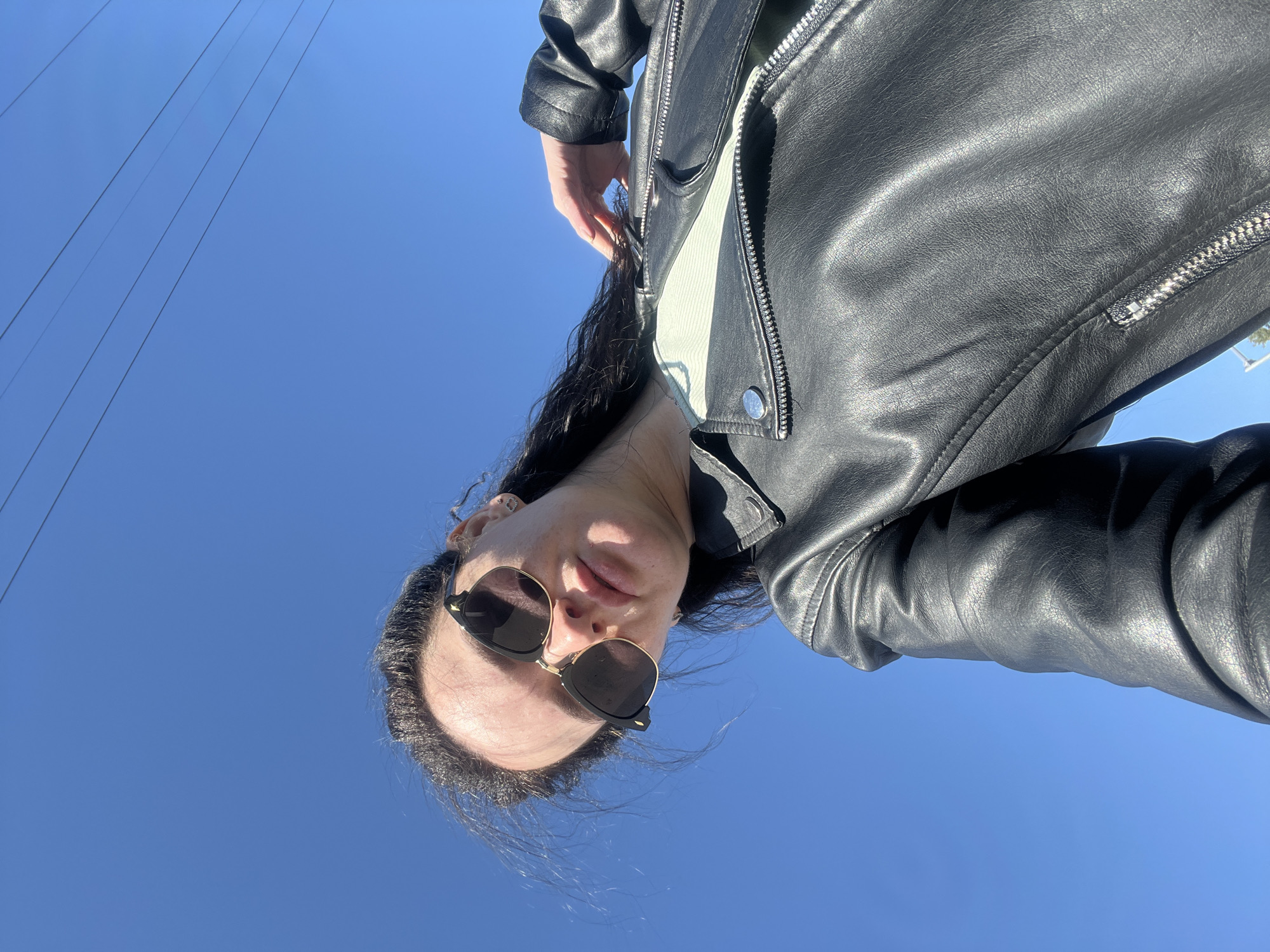 A low-angle, upward-facing photograph depicts a person wearing a black leather jacket and dark sunglasses against a clear blue sky. The individual's face is the primary subject, positioned in the lower portion of the frame. Some power lines are visible in the upper-left. The specific location within Kazan, Russia, cannot be determined from the available visual information.