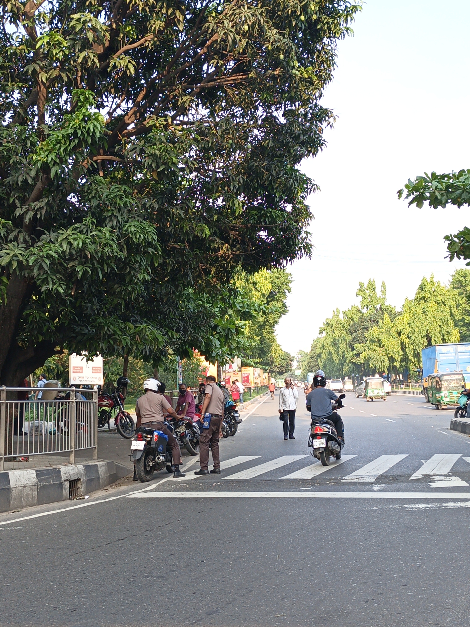 The image depicts a street scene in the city of Unknown, Unknown. The street is asphalt and has a crosswalk in the foreground. Two uniformed officers are standing next to a motorcycle parked on the side of the road. One is on his phone. Another man is walking away from the street. A person is riding a scooter across the crosswalk, heading away from the viewer. There are several other people and vehicles, including auto rickshaws and trucks, on the street. Trees line both sides of the road.