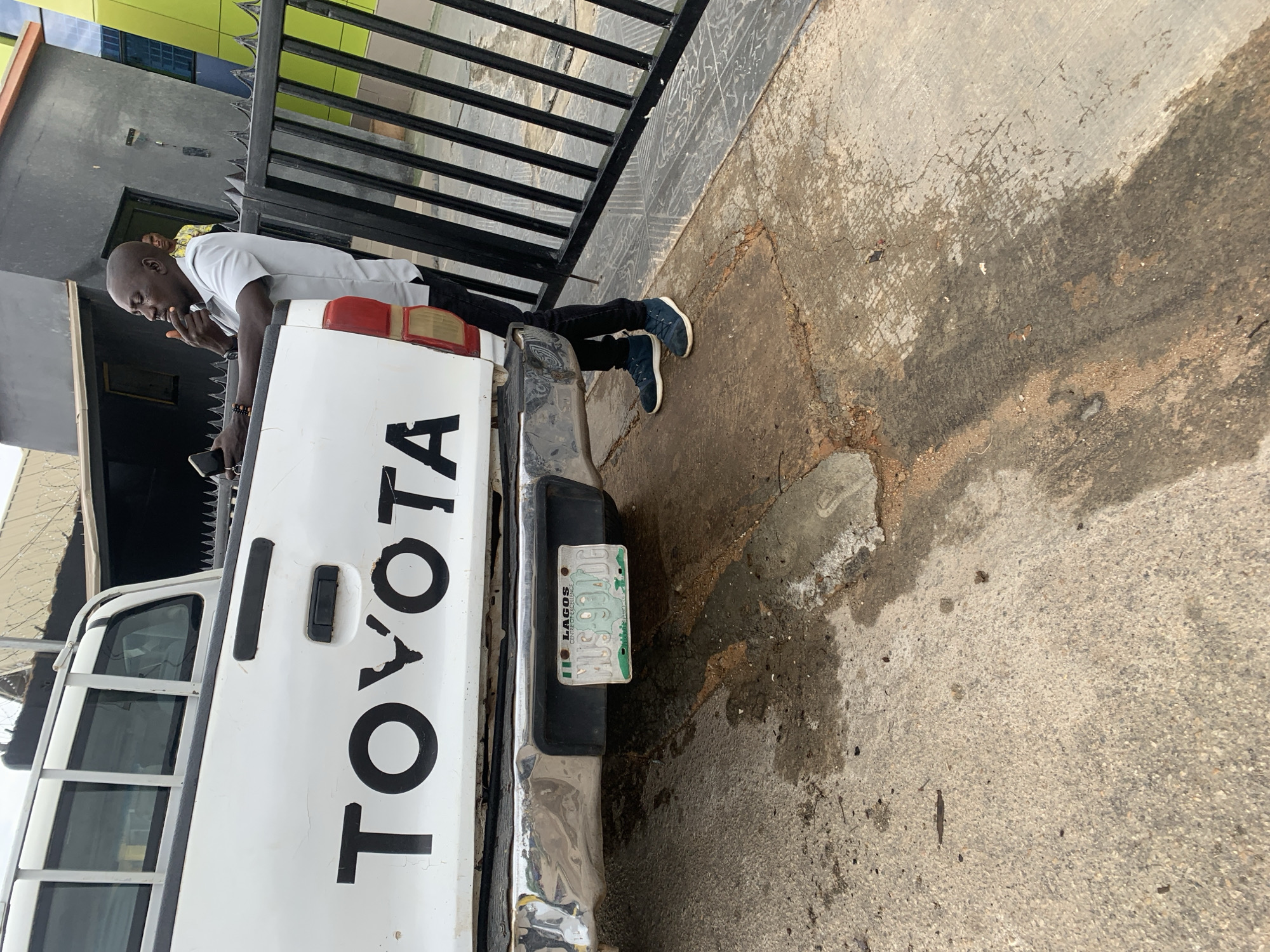 A man stands next to a white Toyota pickup truck with a Lagos, Nigeria license plate that reads "MOS904DG." The man is dark-skinned, bald, wearing a light-colored short-sleeved shirt, dark pants, and dark sneakers. He holds a black object, possibly a phone, near his mouth. The truck is parked next to a black metal fence with a building visible behind the fence. The setting appears to be a paved area, possibly a sidewalk or parking lot. The location is not in Jos, Nigeria, but Lagos, Nigeria as indicated by the license plate.