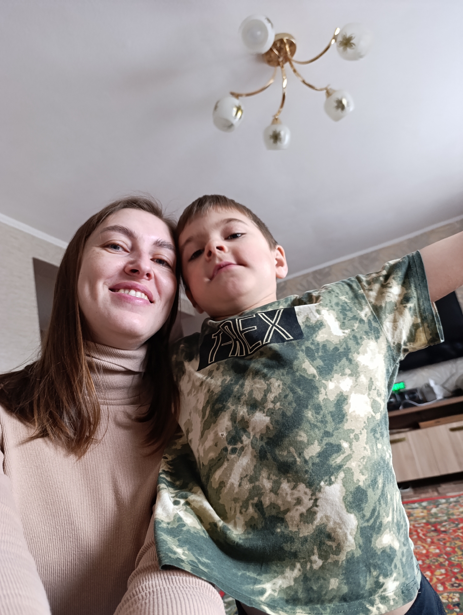A selfie taken indoors shows a woman and a boy, likely in Фоки, Russia. The woman is on the left, wearing a beige turtleneck sweater. She is smiling and looking at the camera. The boy is on the right, wearing a camouflage-patterned t-shirt with the word "REX" printed on it. He also appears to be looking at the camera. In the background, a ceiling with a multi-bulb light fixture is visible. Further back, a television and a wooden cabinet can be seen. A carpet with a patterned design is on the floor.