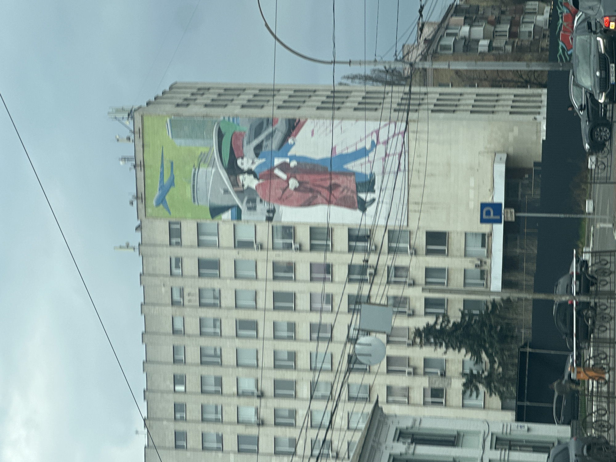 The image depicts a tall, multi-story building in Kyiv, Ukraine, prominently featuring a large mural on its side. The mural portrays a couple in stylized attire, with one figure in red robes and another in blue, set against a backdrop that includes a blue airplane and other abstract elements. The building itself has a grid-like facade with numerous windows and visible air conditioning units. A parking sign is visible near the base of the building. Overhead power lines crisscross the foreground. Parts of the surrounding urban environment, including other buildings and vehicles, are also visible. The sky is overcast.