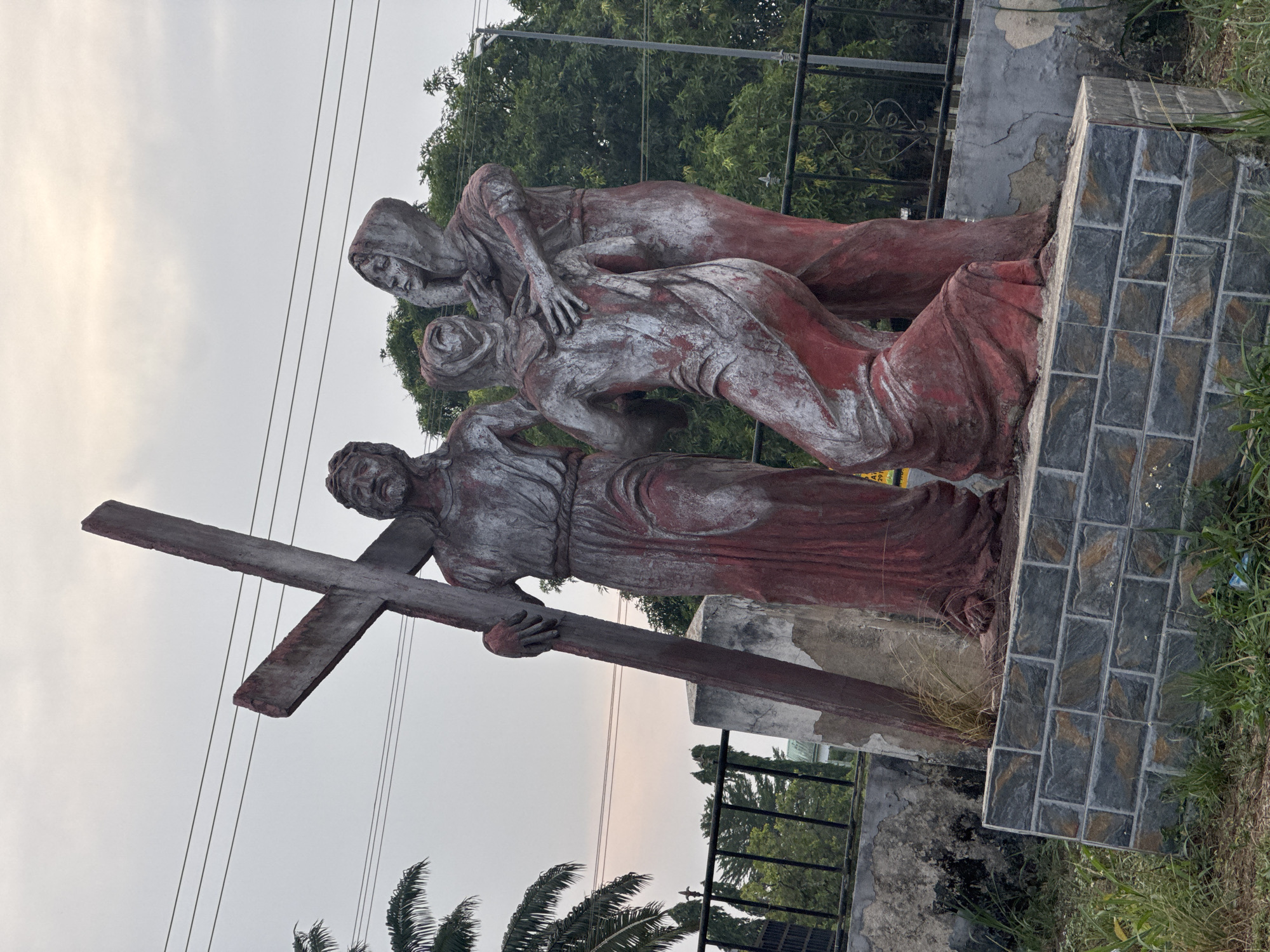 A weathered outdoor sculpture, depicting a segment of the Stations of the Cross, is positioned in Ihiagwa, Nigeria. The scene features three life-sized figures rendered in concrete or similar material, painted primarily reddish-brown with extensive grey and white fading and chipping. A bearded male figure, centrally positioned on the left, bears a large, rough-hewn cross on his right shoulder, gripping it with both hands. His expression is somber. To his immediate right, a figure in a hooded robe kneels, or appears to be collapsing forward, while being embraced and supported from behind by a third standing figure. This third figure, also robed and hooded, places both hands on the kneeling figure's back and shoulder. The entire sculpture group rests on a tiered base. The lower tier is clad with dark grey rectangular tiles arranged in a running bond pattern, exhibiting faint brown and gold streaks. The upper tier is a plain concrete slab, showing signs of wear. The background features an overcast, light grey sky traversed by three parallel overhead power lines. Dense green foliage, including palm trees, forms a backdrop. A black wrought-iron fence and weathered concrete structures are visible behind the sculpture, with sparse dry grass and small plants growing around the base.