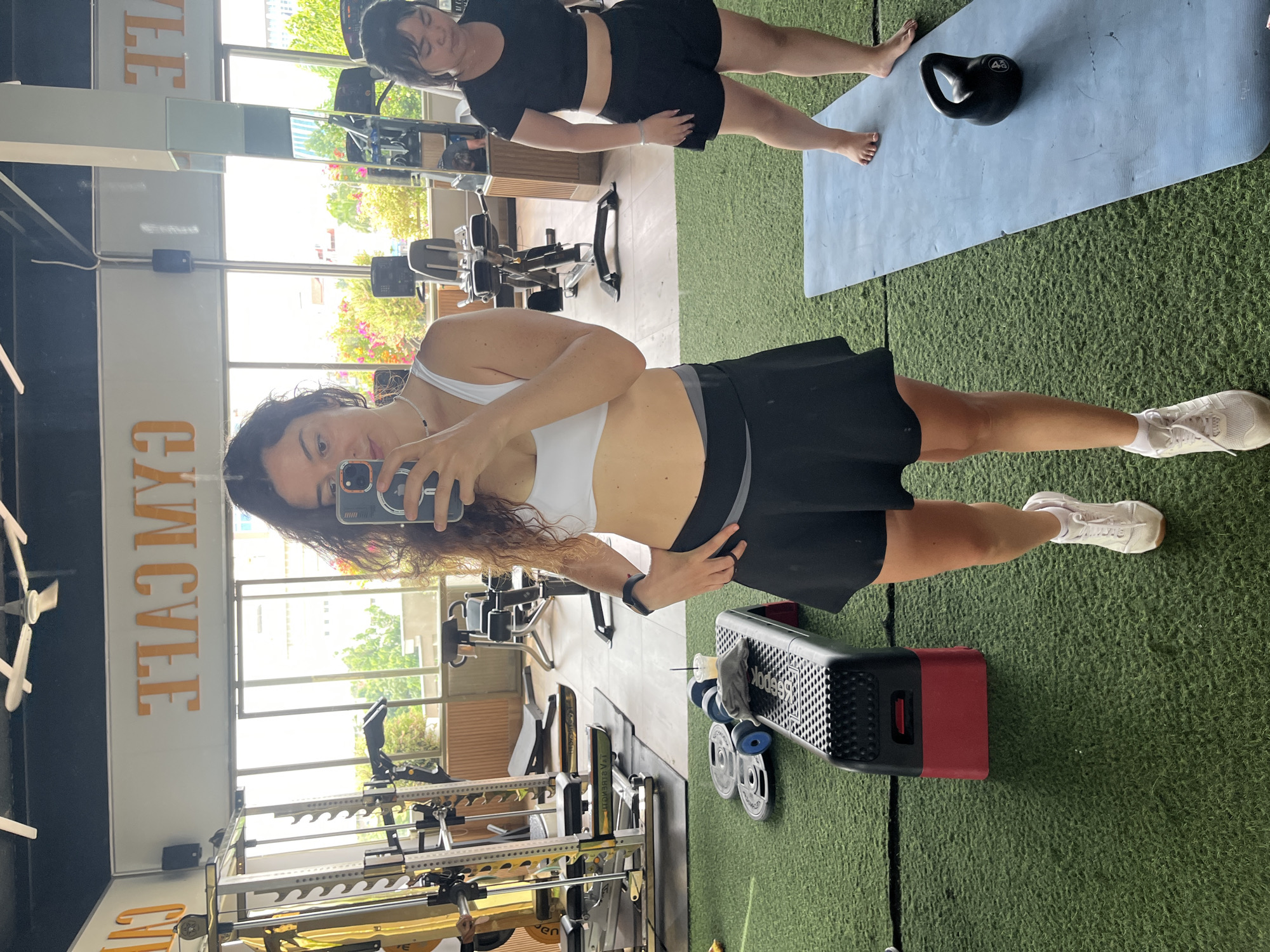 A woman is taking a mirror selfie in a gym. She is wearing a white sports bra and a black skirt. She is standing in front of a large mirror, and her reflection shows her body from the waist up, along with the gym equipment in the background. Another woman is visible in the background, to the right of the main subject, wearing a black sports bra and black shorts. The gym has a green artificial turf floor and is equipped with various exercise machines. The sign above the women reads "GYM CAFE" in orange letters, reflecting in the mirror. The overall environment suggests a well-equipped fitness center with large windows offering a view of the outdoors, indicating it might be located in a city. Specifically, this gym is in the city of Phan Thiết, Vietnam.