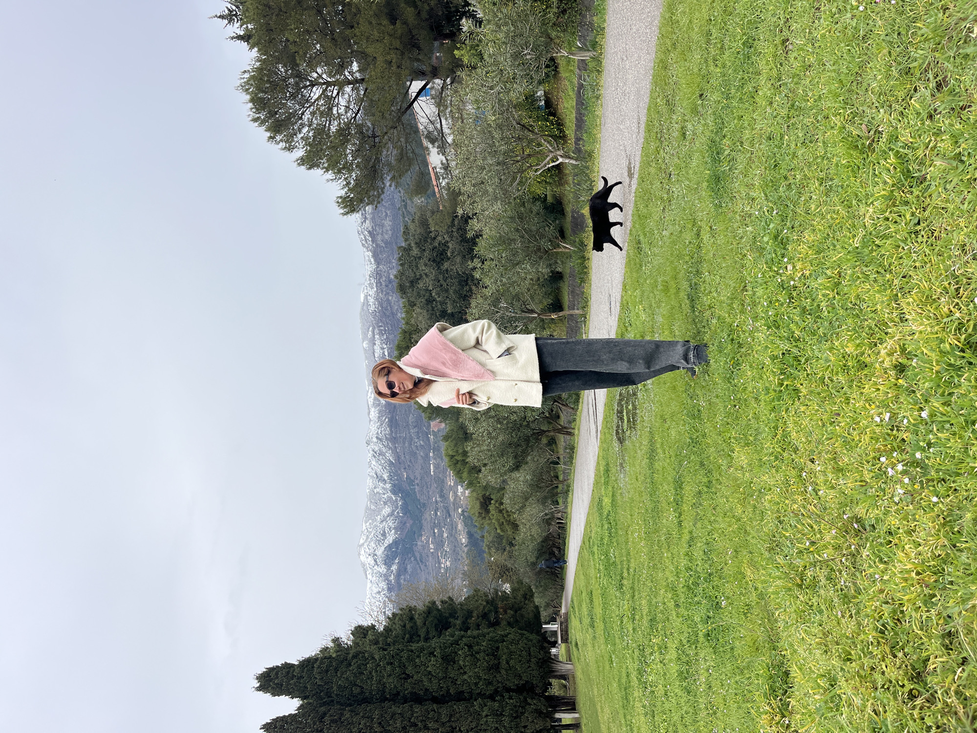 The image depicts an outdoor scene in Budva, Montenegro, under an overcast sky, with snow-capped mountains in the distance.
A female subject stands centrally in the mid-ground on a grassy area. She has reddish-brown hair, wears sunglasses, a cream-colored double-breasted coat, a light pink faux fur scarf, and dark grey wide-leg trousers. She is turned slightly to her right, with a subtle smile. Her left hand is in her coat pocket, and her right hand rests on her scarf.
To the right of the woman, a black cat is captured mid-stride, walking from left to right on a grey paved pathway. A small puddle is visible on the path near the foreground. Further along the path, in the background, a second individual wearing dark clothing walks away from the camera.
The foreground consists of green grass with small white flowers. A row of tall, dark green cypress trees lines the left side of the frame. To the right of the paved path, a line of smaller, light green trees, consistent with olive trees, extends into the background. The horizon is dominated by snow-capped mountains, their lower slopes featuring scattered residential structures and green foliage. The sky is uniformly light grey and overcast.
