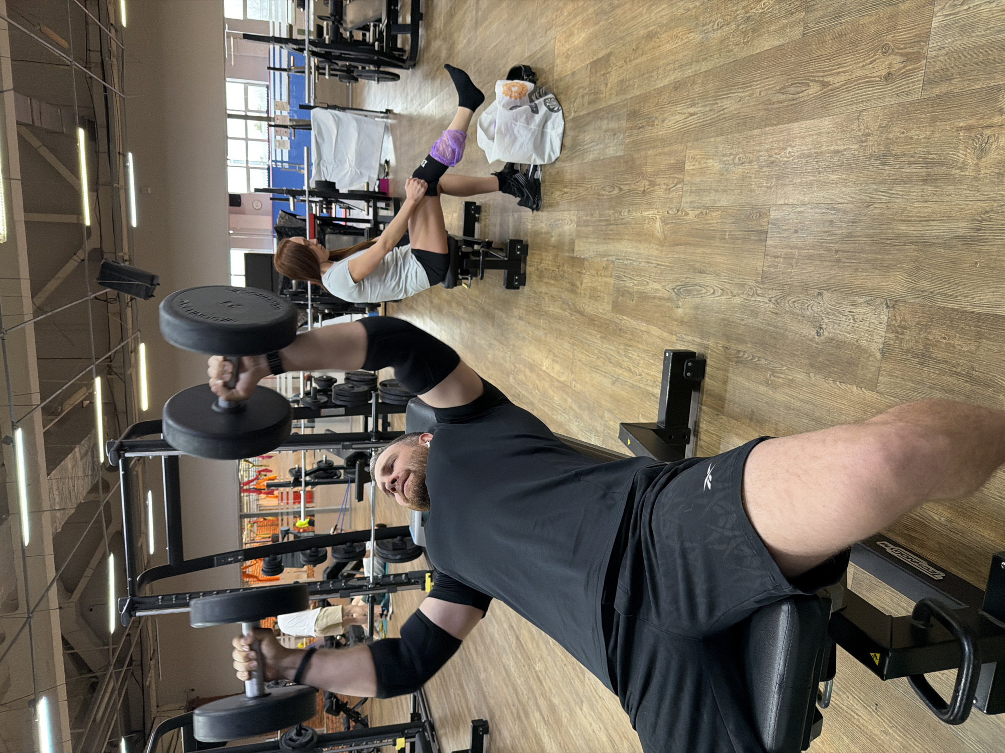 This image captures an indoor gym scene in Chaikovsky, Russia, depicting two individuals engaged in strength training exercises. The photograph is oriented ninety degrees clockwise.
In the foreground, a man with a beard is lying on his back on a black exercise bench, holding a heavy black dumbbell in each hand, extended upwards as if performing a chest press or fly. He is wearing a black t-shirt, black shorts with a visible "REEBOK" logo, and black elbow sleeves. He looks towards the camera with a slight smile. The bench he is using has the brand name "TECHNOGYM" printed on it.
In the background, a woman with long brown hair is also exercising, positioned on a different piece of gym equipment. She is lying with her legs bent and raised, wearing a light-colored t-shirt, black shorts, black socks, and black athletic shoes. A distinctive purple support or wrap is visible on her left lower leg. Near her feet, on the wooden floor, there is a white bag and several water bottles or cups.
The gym environment features a light brown wooden floor with visible grain, and a high ceiling equipped with multiple fluorescent strip lights and exposed structural elements. Large windows are visible in the background, suggesting natural light is also entering the facility during the daytime. Various other weight racks, exercise machines, and gym equipment are scattered throughout the spacious area. The dumbbells held by the man have numbers embossed, with "15" possibly visible on one of them.