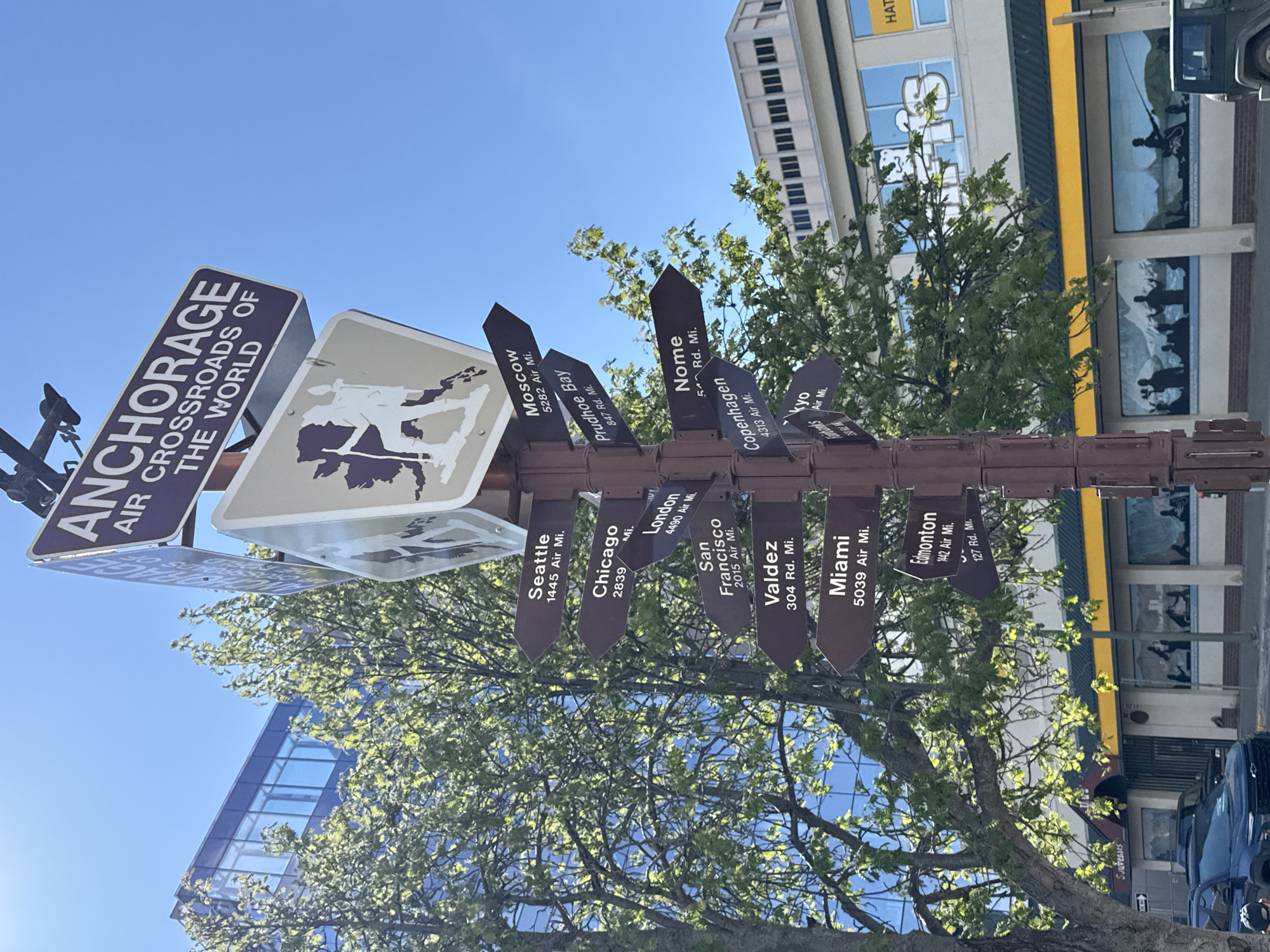 The image shows a signpost in Anchorage, Alaska, United States, indicating various cities and their distances. The main sign reads "Anchorage Air Crossroads of the World." Attached to the post are several arrow-shaped signs pointing in different directions with the names of the cities and their distances, including Seattle (1445 Air Mi.), Moscow (5282 Air Mi.), Prudhoe Bay (847 Rd. Mi.), Chicago (2839 Mi.), London (4490 Air Mi.), San Francisco (2015 Air Mi.), Valdez (304 Rd. Mi.), Miami (5039 Air Mi.), Nome (5 Rd. Mi.), Copenhagen (4313 Air Mi.), Edmonton (1442 Air Mi., 127 Rd. Mi.) and Tokyo. The signpost is surrounded by trees with green leaves, and behind it are buildings. A light blue sky is visible at the top of the frame. A building with murals is in the background to the right. A vehicle is visible to the right.