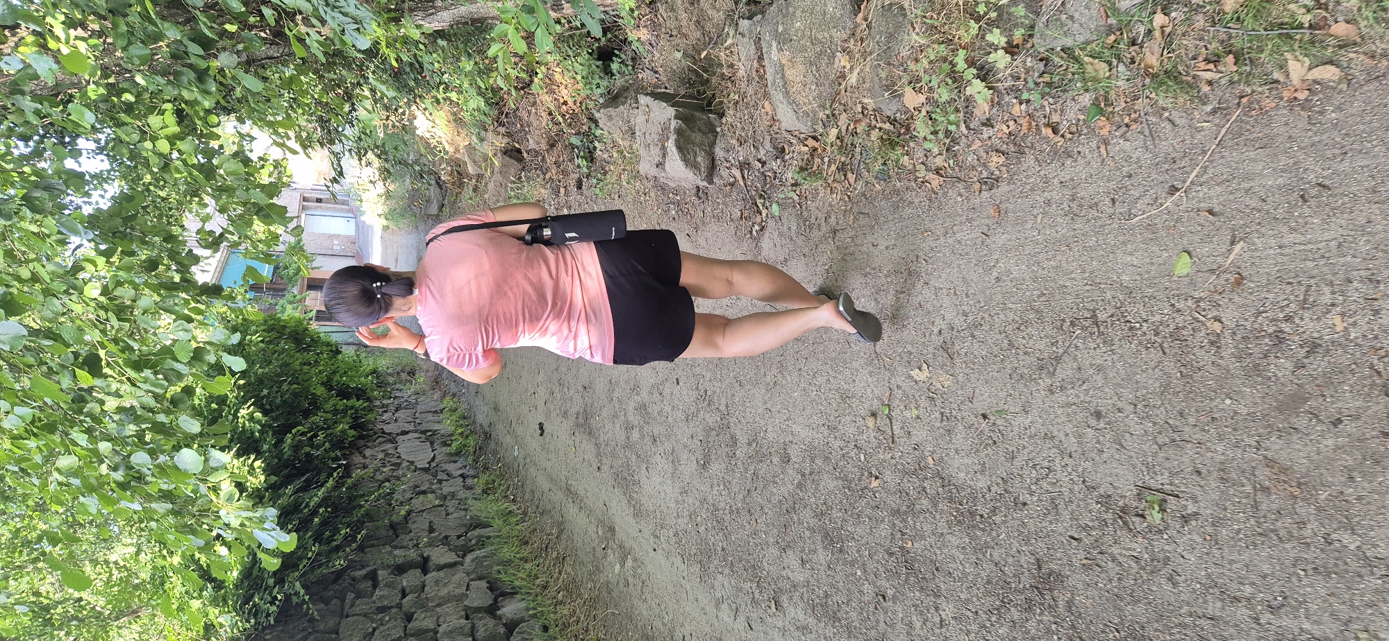 A woman with dark hair pulled into a ponytail walks away from the camera on a dirt path. She is wearing a short-sleeved pink t-shirt and black shorts. A black water bottle with a white logo is slung over her right shoulder. She is wearing dark flip-flops. The path is lined with a stone wall on the left and trees and foliage on the right. Buildings with blue awnings are visible in the distance at the end of the path.