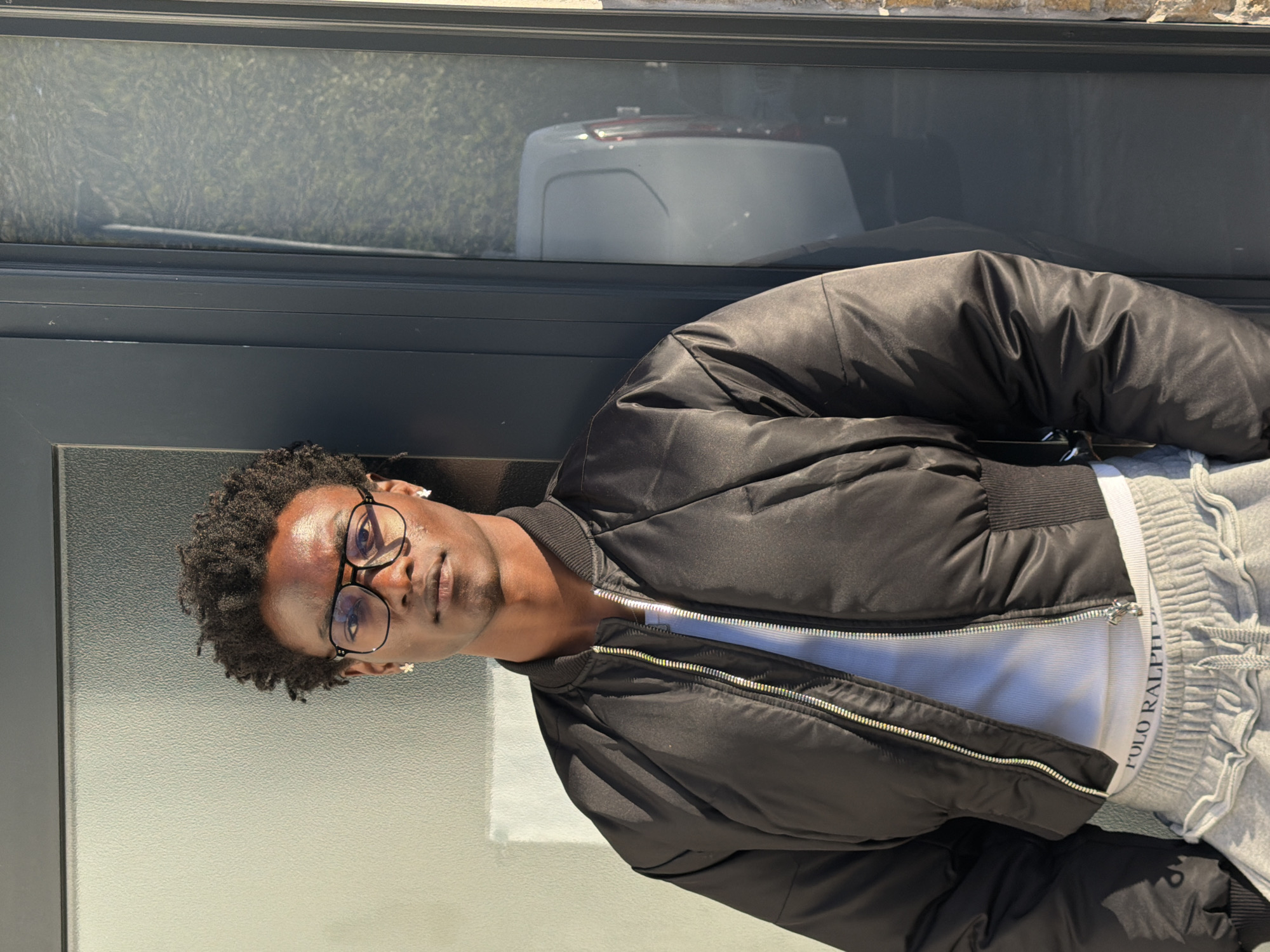 A young Black man with short, curly dark hair stands and looks directly at the camera. He wears dark-rimmed glasses and a small, white, possibly floral, earring in his left ear. He is dressed in a black, shiny bomber jacket with silver zippers, worn open over a white ribbed t-shirt. Grey sweatpants are visible below the jacket, with a white waistband displaying the text "POLO RALPH".
The man is positioned outdoors or by a large window/glass door, leaning against a dark-framed structure. Behind him, the reflective surface of the glass shows a blurred reflection of a bright, sunny environment. A distinct white object, resembling the rear of a vehicle (possibly a car or van), and some green, textured elements suggestive of foliage or a distant building are visible in the reflection. The scene is brightly lit by natural sunlight, indicating it is daytime. The man maintains a neutral expression.