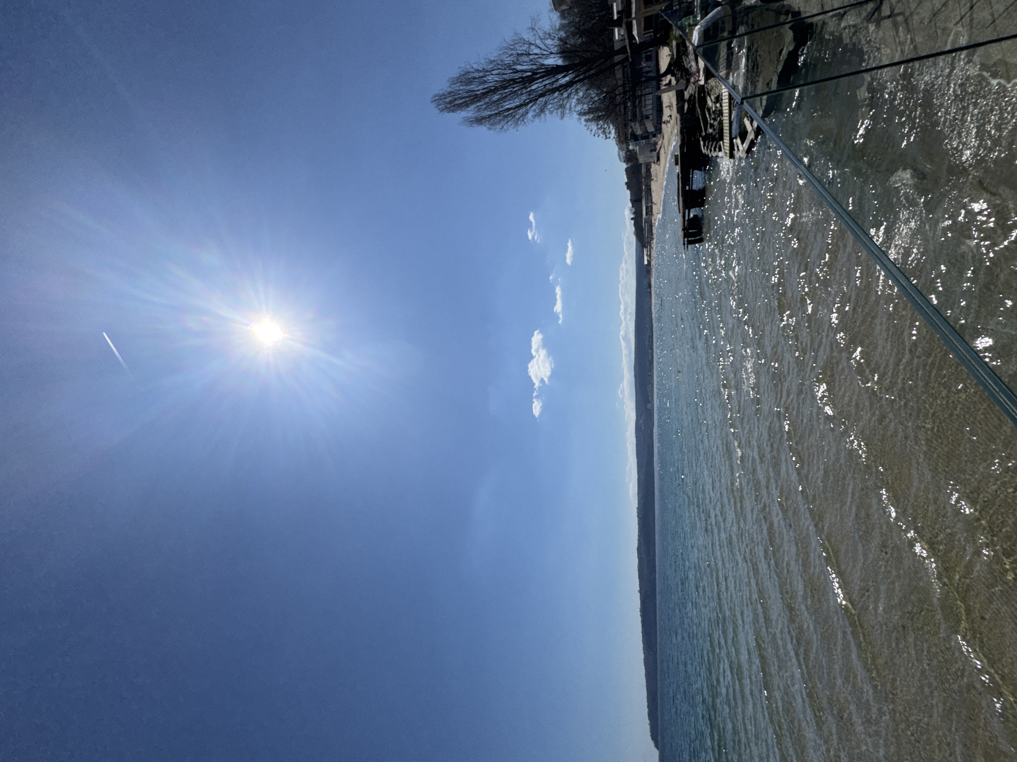 The image depicts a sunlit scene in Varna, Bulgaria. It showcases the Black Sea and a portion of the coastal area. The blue sky occupies a significant portion of the image, with the sun emitting strong rays. A few small clouds are scattered in the sky. The calm sea reflects sunlight, creating a shimmering effect. Along the shoreline, there are buildings and trees. A glass barrier is visible, suggesting an elevated perspective or a balcony setting.