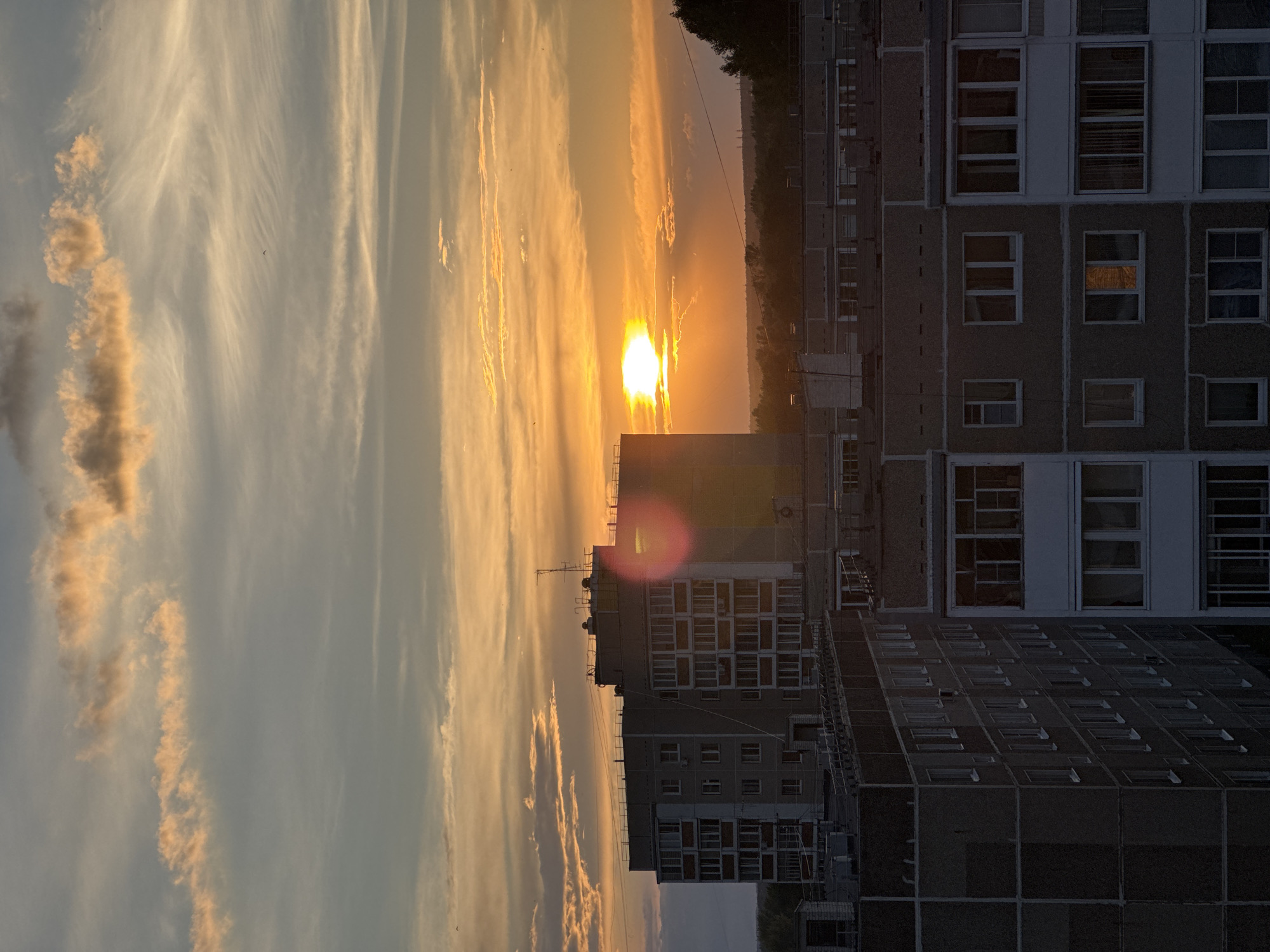 This media file depicts a sunset over a residential area in Chaikovsky, Russia. The image is oriented vertically, showing a vast sky filled with wispy clouds tinged with the golden hues of the setting sun. A bright, prominent sun is visible near the center of the frame, casting a warm glow and creating lens flare. Below the sky, the silhouettes of several apartment buildings dominate the lower portion of the image. These buildings are characterized by their repetitive window patterns, typical of Soviet-era architecture. Some windows reflect the sunset's light, while others appear dark. In the distance, a line of trees marks the horizon, and faint industrial structures can be seen. The scene evokes a sense of twilight and the end of the day in an urban environment. The weather appears clear with scattered clouds. There is no visible text in the image.