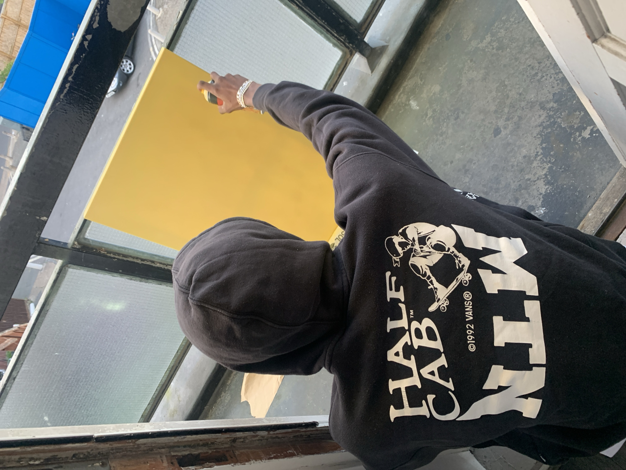A person wearing a black hoodie with the Vans Half Cab logo on the back is applying yellow paint to a surface. The person is wearing a silver bracelet on their left wrist. The location appears to be outdoors, possibly a building exterior or balcony. The environment includes a window frame and a view of a street with cars. The scene might be in London, United Kingdom, given the urban setting and architectural style.