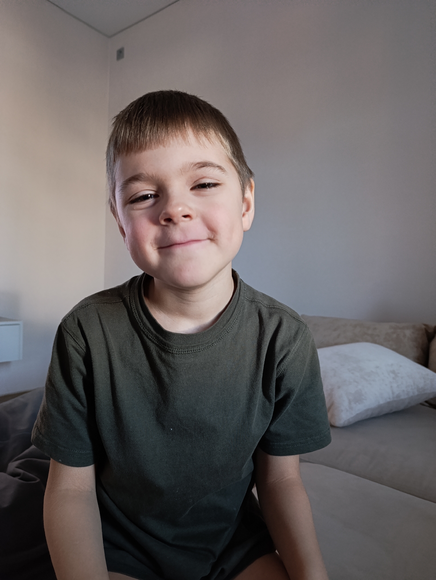 A young boy with fair skin, short brown hair, and bangs is captured in a head-and-shoulders portrait, looking directly at the camera with a gentle, closed-lip smile. His dark eyes are open, and faint dimples are visible on his cheeks. He is wearing a dark olive green, short-sleeved t-shirt.
The boy is seated indoors, likely on a bed or sofa, with a light brown or beige upholstered headboard or backrest visible behind him to the right. A white pillow rests against this backrest. The background features plain light beige walls, with a corner visible in the upper left, suggesting a simple room interior. A small, white, wall-mounted shelf or cabinet is partially visible on the far left. The lighting is soft and natural, suggesting it is daytime. The boy's expression conveys contentment and happiness. No visible text is present in the image. The setting is Chaikovsky, Russia.