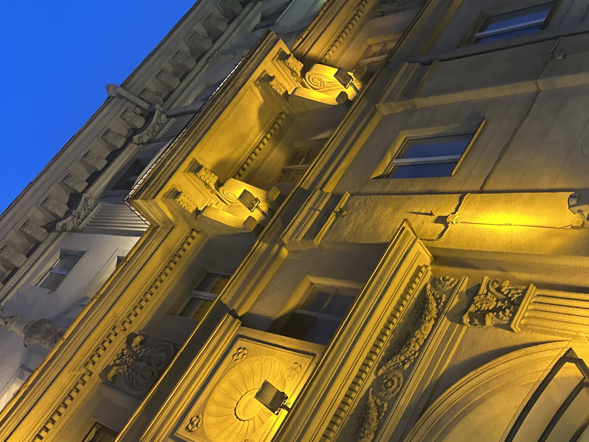 This image depicts a building facade in Minsk, Belarus, illuminated by warm yellow artificial lights against a deep blue twilight sky. The architecture is ornate, with classical elements such as columns, decorative moldings, and sculpted reliefs visible on the building's exterior. Windows with modern frames are visible, some reflecting the sky. The lighting emphasizes the architectural details and casts shadows, creating a dramatic effect. There are no people or active events depicted in the scene. The image focuses solely on the static architectural features of the building.