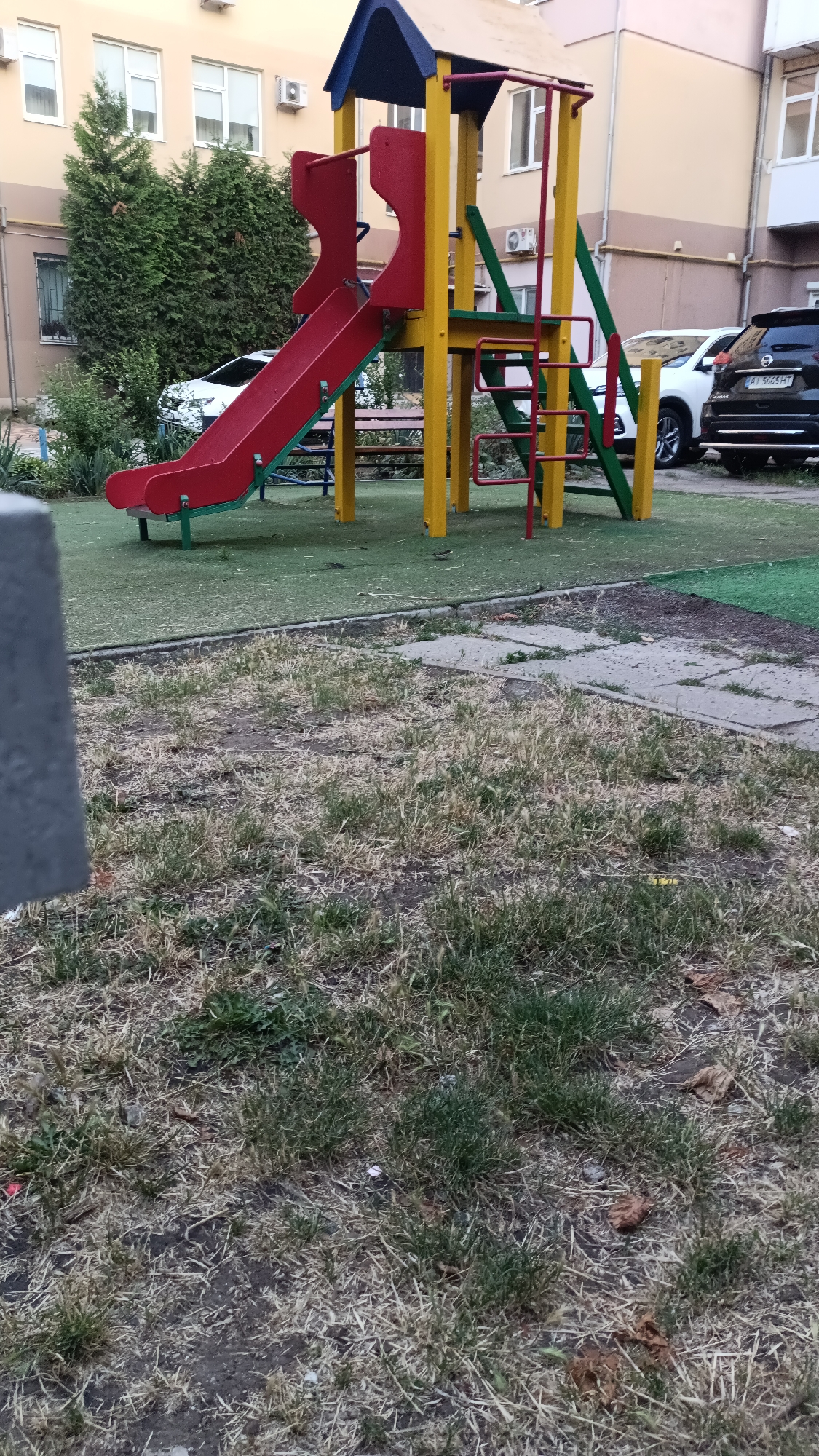 The image depicts a children's playground in the city of Chornomorsk, Ukraine. The playground features a red and yellow slide structure with a blue roof. A red slide descends from a platform, supported by yellow and green vertical poles. A red ladder with green rungs leads up to the platform. The playground surface appears to be green artificial turf. Behind the playground, there is a light-colored building with windows and air conditioning units. Two cars are parked near the building: a white SUV and a dark SUV with license plate AT 5665 HT. The foreground of the image shows dry, patchy grass and concrete paving stones.