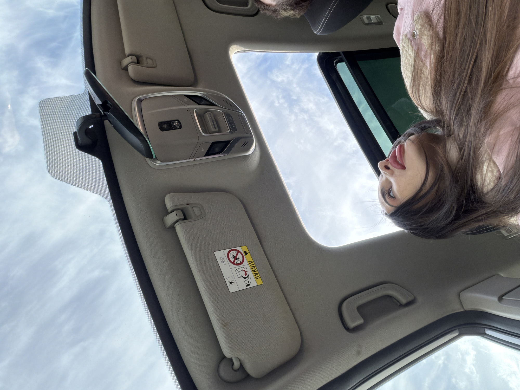The image shows the interior of a car, with the ceiling, sun visor, rearview mirror, sunroof, and a portion of a person. The person is leaning into the car, with their head and shoulders visible. The sky with scattered clouds is visible through the sunroof. No specific location in Saint Petersburg, Russia, is identifiable from the image.