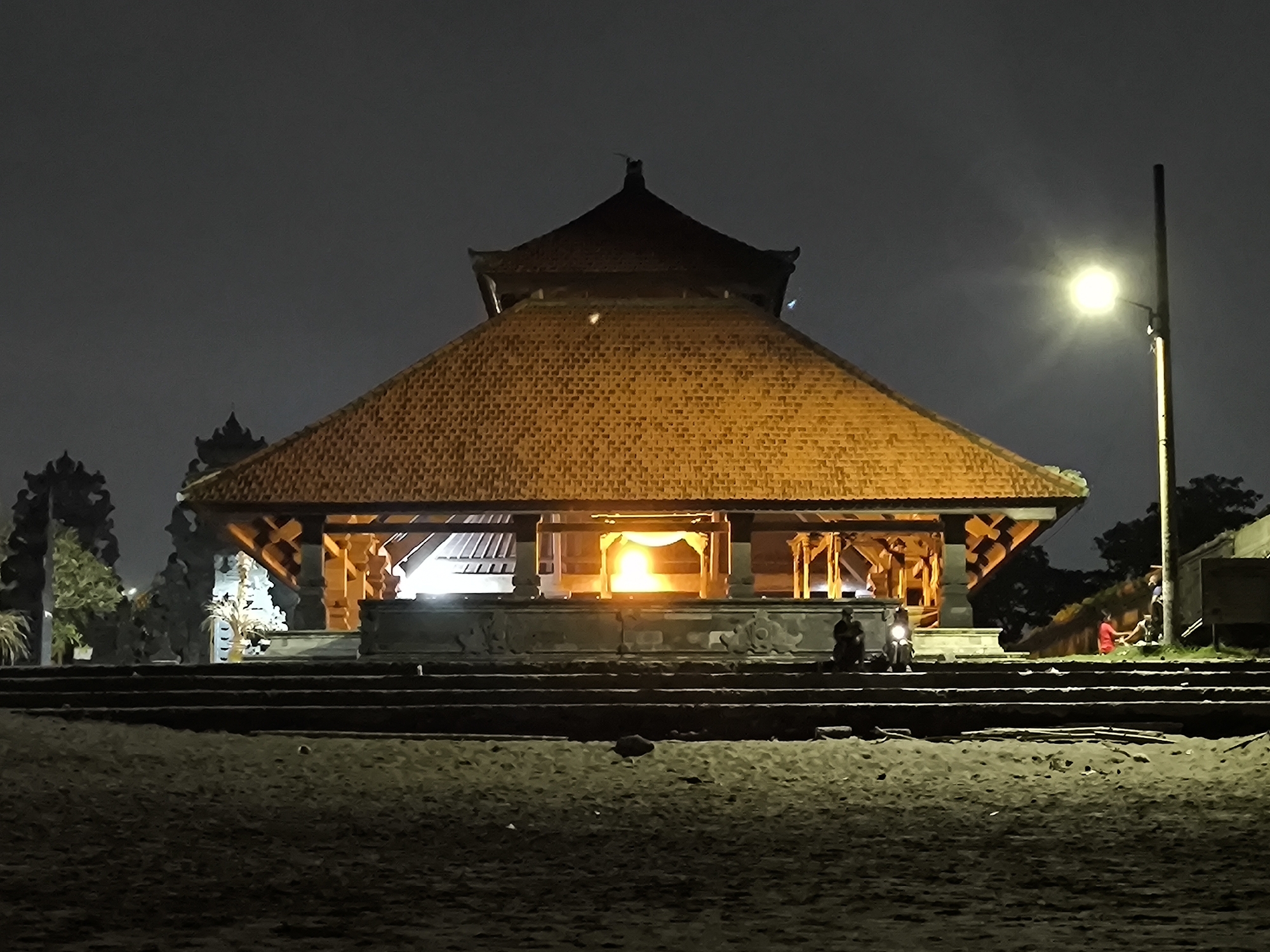 At night in Canggu, Indonesia, a large Balinese temple structure with a multi-tiered tiled roof is illuminated. The structure is set back from the viewer and sits atop a series of stairs. A bright light emanates from the interior, contrasting with the surrounding darkness. A street lamp stands to the right of the temple. Several people are present; some are seated on the steps near the temple, while others are gathered nearby. The foreground is an open, dark area, appearing to be a sandy or grassy expanse.