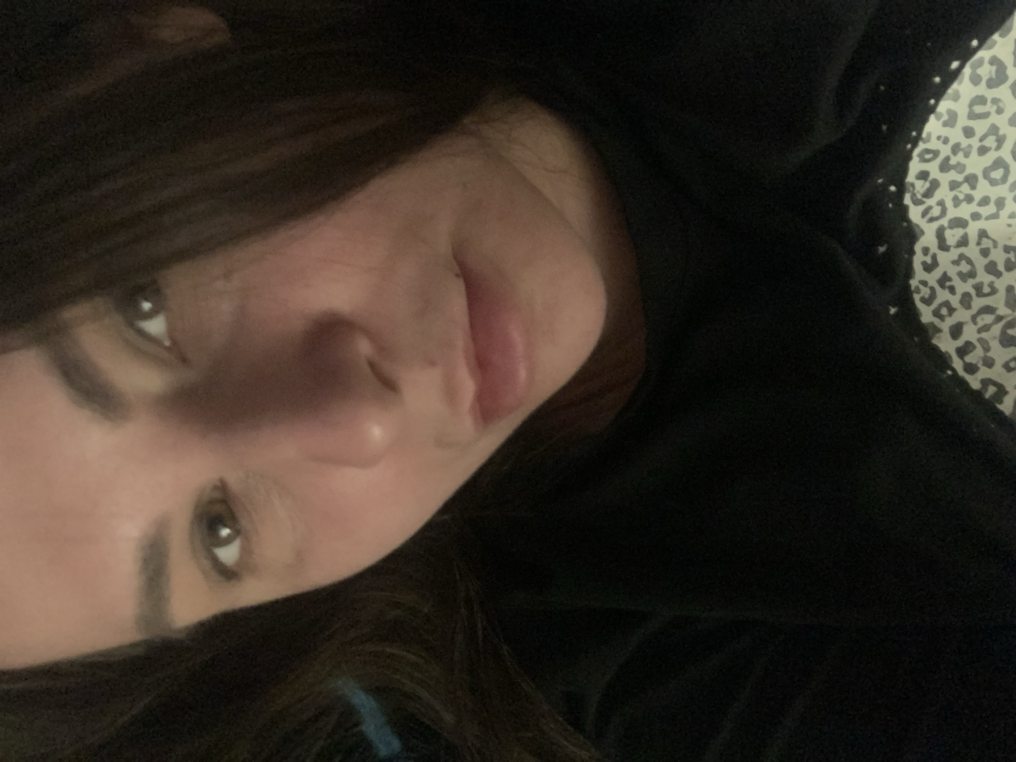 A close-up, angled, selfie-style shot captures the face of a young person with brown hair and brown eyes. They are wearing a black garment that appears to be a shirt or sweater, which is partially visible in the upper portion of the frame. The background behind the person's head features a leopard print pattern, suggesting they are lying on a surface with this design. The lighting is soft and diffused, with some highlights on the face. The image is oriented such that the person's face is upside down.