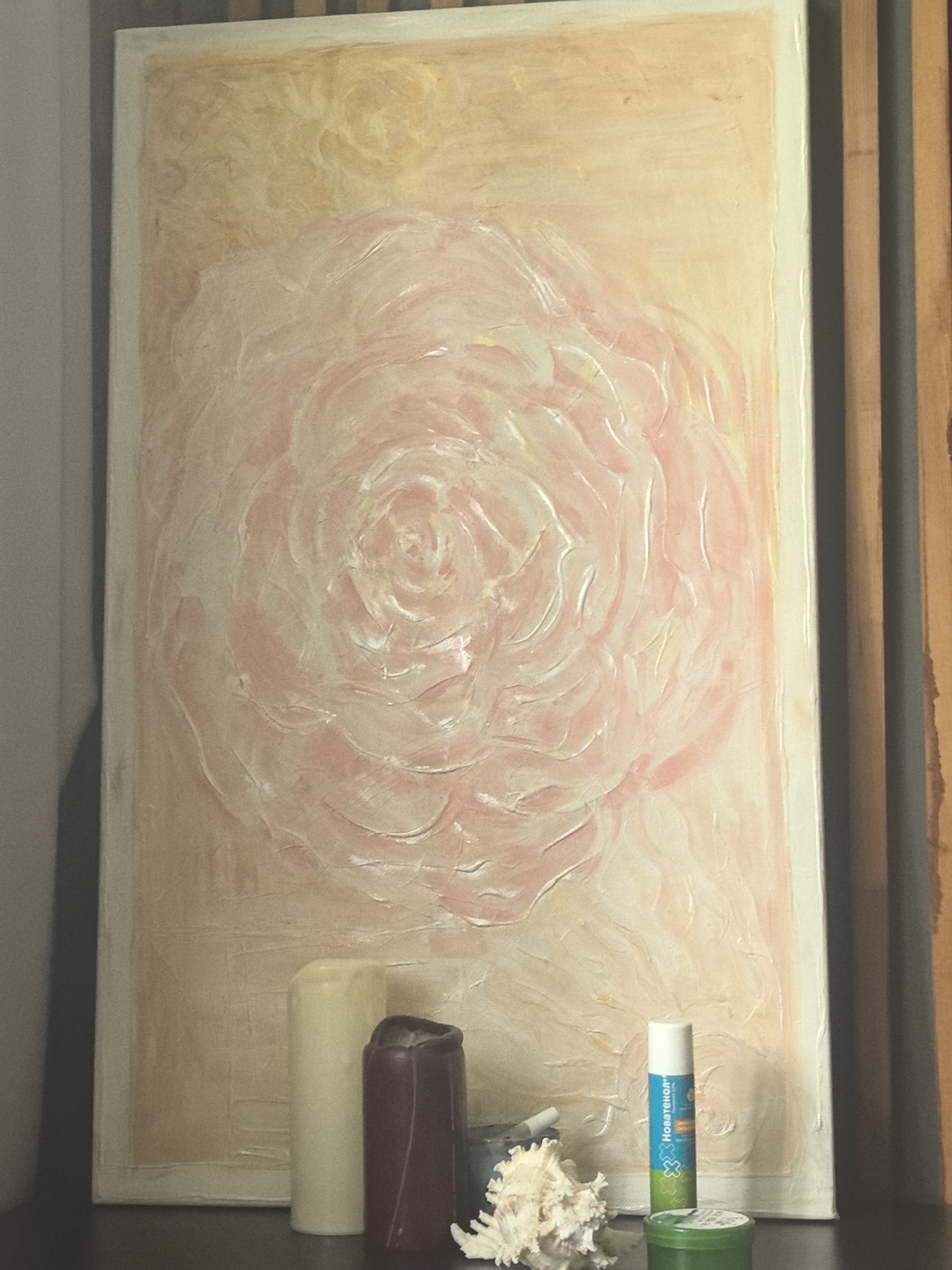 The image shows a painting of a pink and white rose, textured with thick brushstrokes. The painting is framed with a white border and leans against a wall. In front of the painting, on a dark surface, are two candles, one cream-colored and the other dark purple. To the right of the candles is a small seashell and a tube of lip balm. The background consists of a wall with vertical wooden slats on the right side. The scene is dimly lit, casting shadows. There are no people visible in the image. The location of this scene in the city of Khabarovsk, Russia cannot be determined from the image itself.