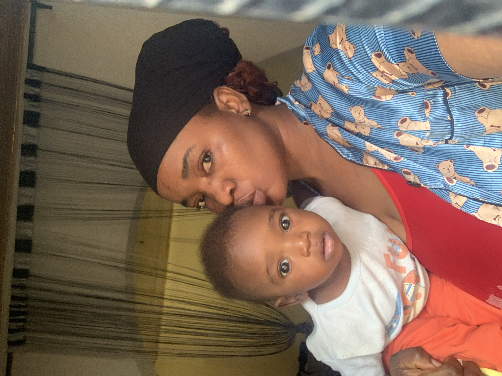 A vertical portrait-style photograph, likely taken indoors in Barakin Akawo, Nigeria, captures a tender moment between a woman and a baby. The image is rotated, presenting the subjects as if positioned side-by-side with the woman on the left and the baby on the right.
The woman, with dark skin, wears a black head wrap that covers most of her hair, though some reddish-brown curls are visible near her left ear, which features a small silver earring. She is dressed in a vibrant blue and white striped top patterned with numerous light brown teddy bears, worn over a red undershirt or tank top. Her gaze is directed towards the camera, while her head is gently tilted down, suggesting she is kissing the baby's forehead or temple.
The baby, appearing to be a toddler, is held close by the woman. The baby has dark skin, short, curly dark hair, and large, expressive dark eyes that look directly at the viewer. The baby's mouth is slightly open. Dressed in a white t-shirt with faded, colorful graphics that are not clearly discernible, and orange bottoms, the baby leans into the woman's embrace.
The background features light-colored walls and a prominent pattern of vertical blinds or string curtains, indicating an indoor setting. The lighting is soft and even, highlighting the affectionate connection between the two individuals. There is no discernible text visible on their clothing or in the background elements. The overall mood of the image is warm and loving.