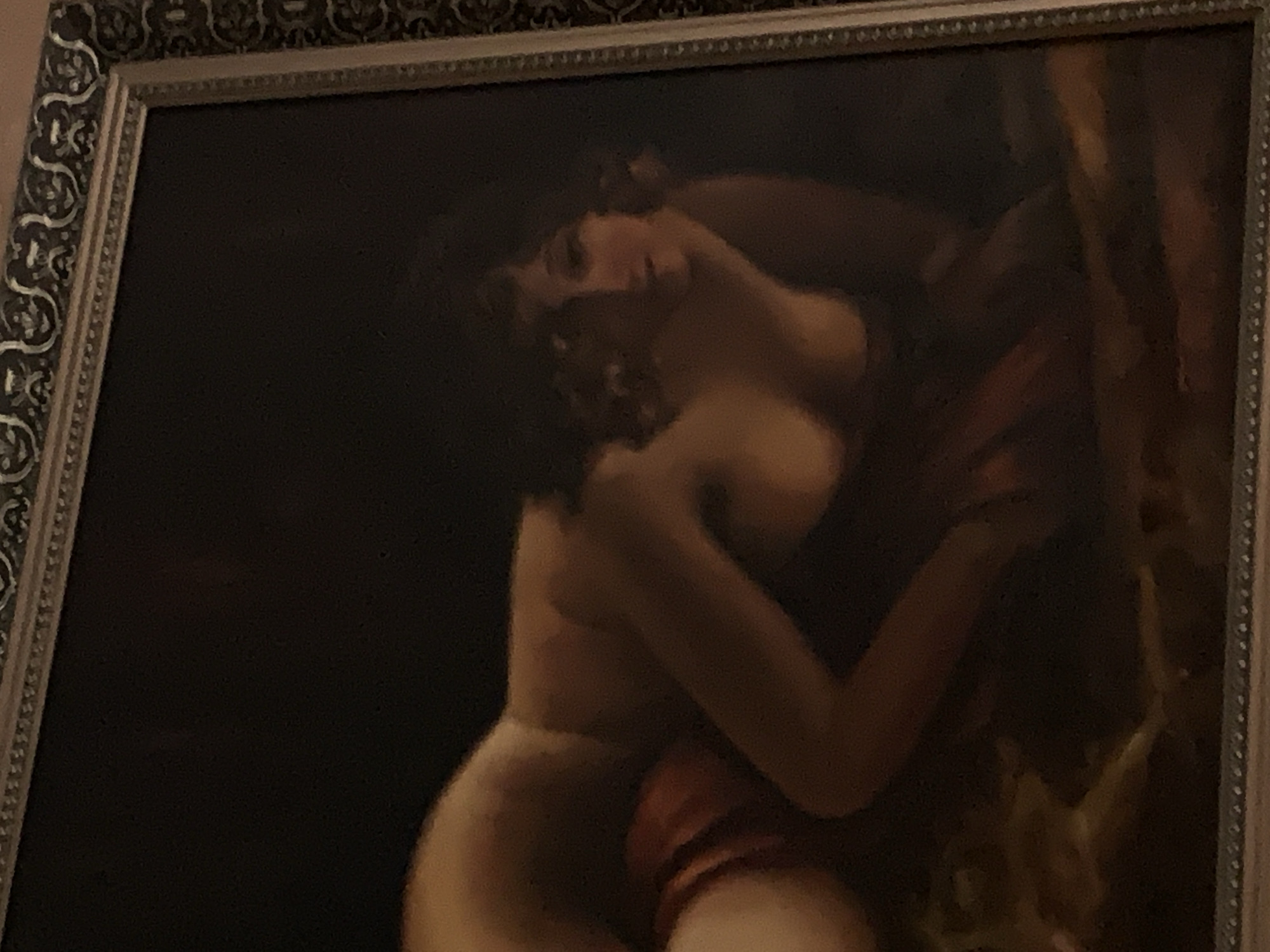 The image is a painting framed by an ornate silver border. The painting itself is dark and somewhat blurry, depicting two figures intertwined. The foreground figure appears to be a woman with her back to the viewer, her body partially obscured by fabric. Her skin tone is light, and she has dark, curly hair. Her head is turned, and she is looking upwards.
Behind her, another figure is visible, embracing her. This figure is darker, and their features are less discernible. The embrace suggests intimacy or passion. The figures are positioned against a dark background, which offers no specific clues about the setting or time of day. The overall mood of the painting seems to be one of intense emotion, possibly romantic or dramatic, conveyed through the close physical contact and the dramatic lighting that highlights the figures. There is no visible text in the image.