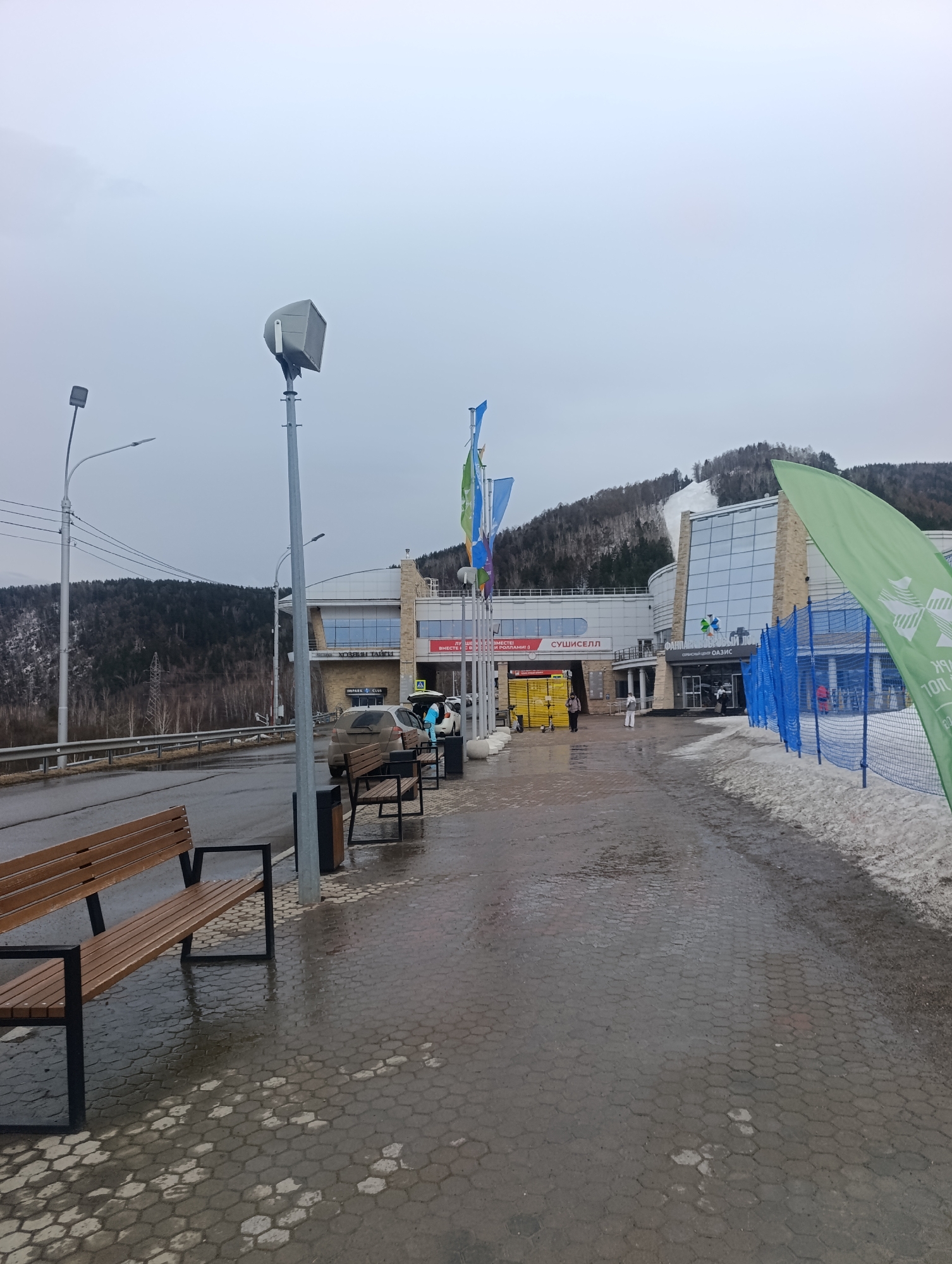The image shows a wet, paved area in front of a building complex with a partial view of a snowy ski slope on the hillside in the background, taken during a cloudy day. A wooden bench sits on the left foreground. A street lamp and a metal waste receptacle stand nearby. Cars are parked near the building. A few pedestrians are walking towards the entrance of the building, which has signs in Russian, including "Сушиселл". Blue fencing and green flags are on the right. The location is likely in or near Krasnoyarsk, Russia, given the signs and architecture.