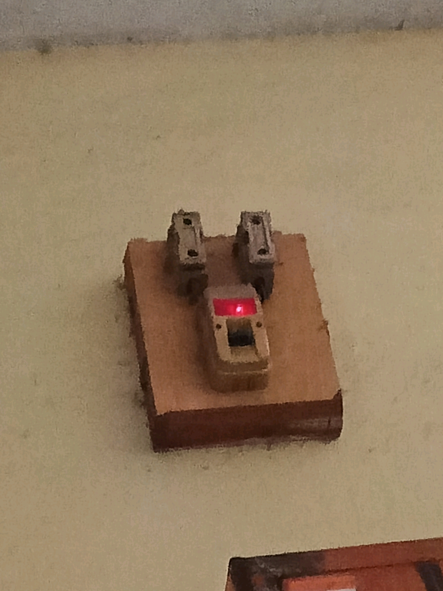 The image shows a close-up of an old electrical fuse box mounted on a beige-colored wall. The fuse box is rectangular and appears to be made of wood, with a weathered texture. On top of the wooden base, there are two rectangular ceramic fuse holders, positioned vertically and spaced apart. A single, red indicator light is visible, glowing brightly on a small, beige-colored component positioned between the two fuse holders. The wall itself has some minor discoloration and texture variations, suggesting it is an interior wall. There are no people or other objects clearly visible in the frame. The overall impression is of a functional, albeit aged, piece of electrical equipment.