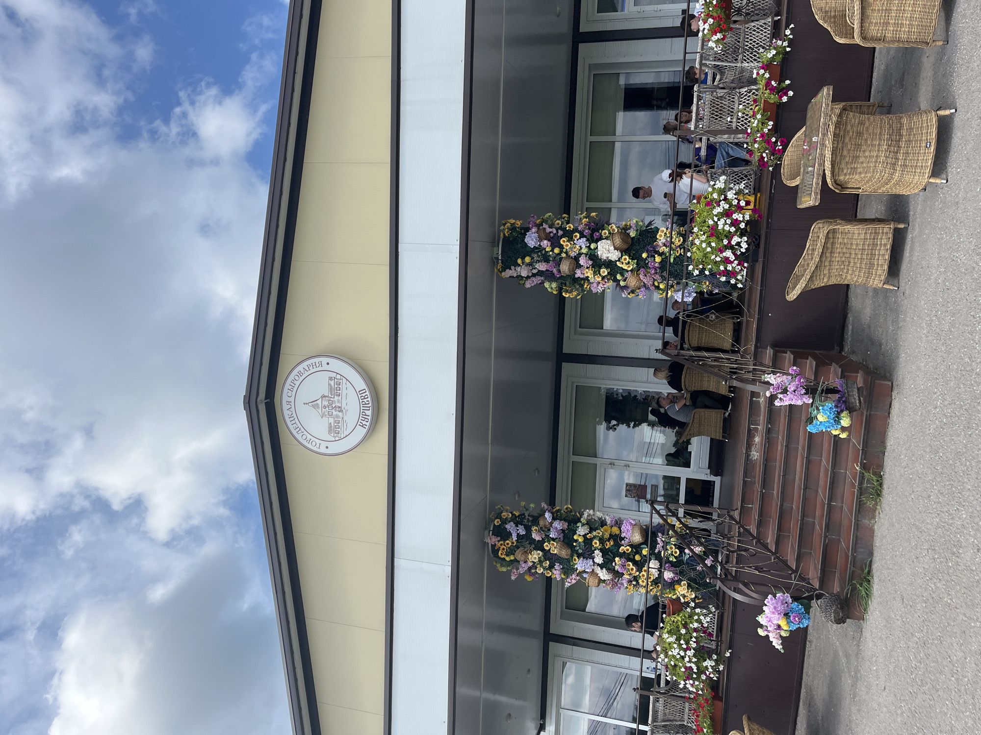 The image shows the exterior of a building, likely a restaurant or cafe, with a sign indicating "GORODETSKAYA SYROVARNYA KURYKHIVO" (Gorodetskaya Cheese Factory Kurikhivo). The building is light yellow with a dark brown roofline. Two large, decorative floral arrangements shaped like columns are placed on either side of the entrance. A set of brick stairs leads up to a covered patio area where several people are seated at tables, some engaged in conversation. The patio is adorned with hanging plants and flower baskets. Wicker chairs and tables are also visible on the patio and in front of the building. The overall setting suggests a pleasant outdoor dining or social gathering space. The sky is cloudy with patches of blue. The location is identified as Akhlebaikha (Kovriginskii S/S), Russia.