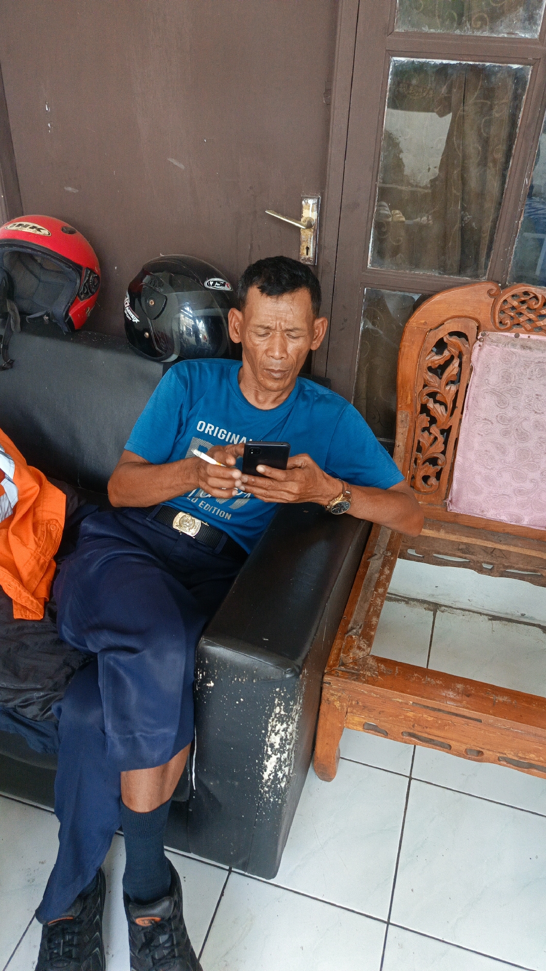 In Margajaya, Indonesia, a man is seated on a dark couch, engrossed in his smartphone. He wears a blue t-shirt, dark trousers, and black socks and shoes. A gold watch adorns his left wrist. Next to him on the couch are two helmets, one red and one black. To his right, a wooden chair with ornate carvings and a pink upholstered seat is visible. The scene appears to be outdoors, possibly on a porch or patio, with a brown door and a glass-paneled door in the background. The floor is tiled in a light color.