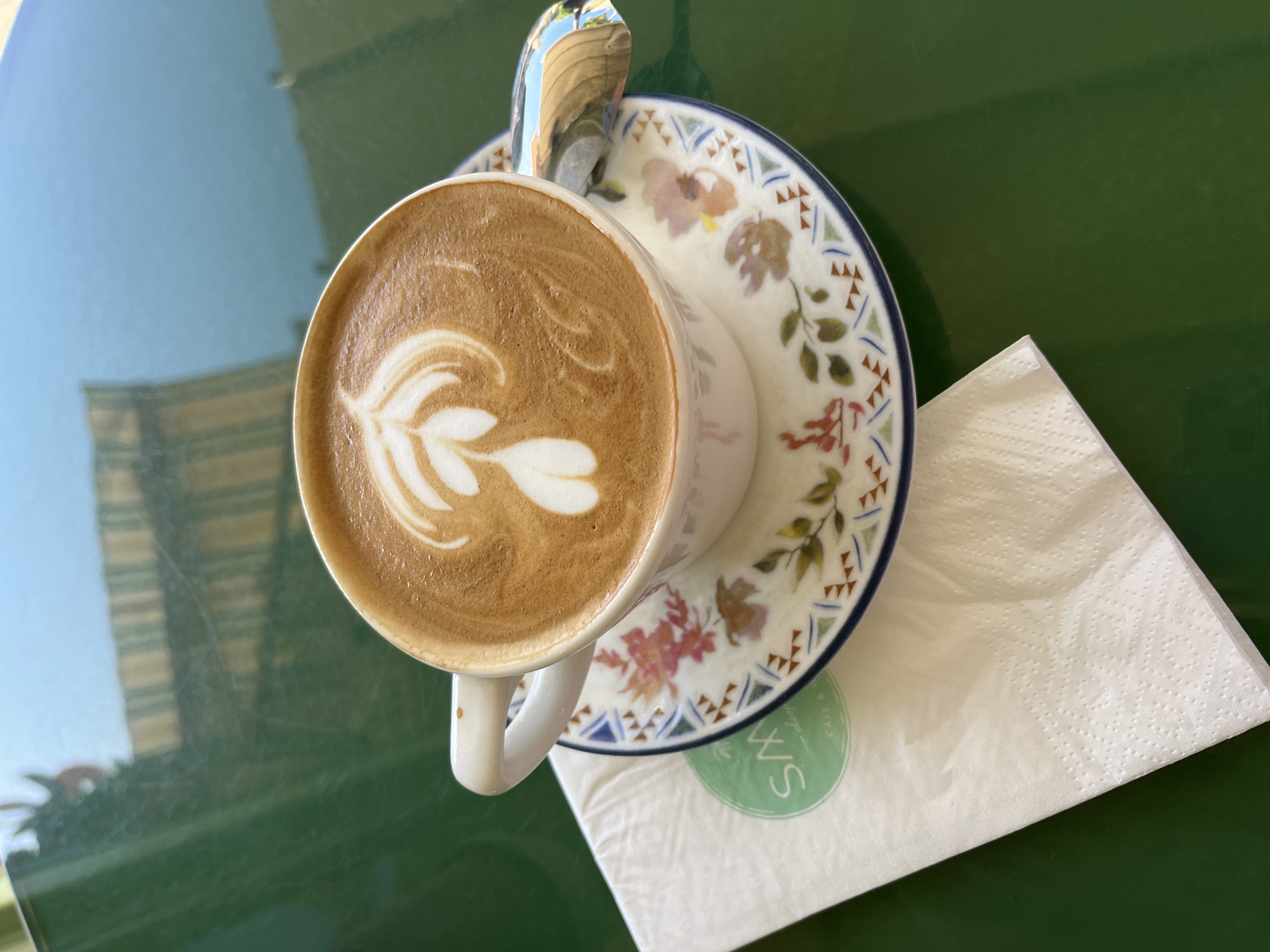 A high-angle shot reveals a cup of latte art placed on a decorative saucer with floral designs. A spoon rests within the cup, partly visible above the latte's surface. Adjacent to the saucer is a white napkin, bearing a circular logo with the initials "S&W" and the words "CAFE & LOUNGE", indicating it may be the cafe where this photo was taken. The latte cup and saucer are positioned on a green, reflective table surface, suggesting an outdoor setting with the reflection of nearby buildings. Given the cafe’s branding and the overall setting, the location is most likely Alanya, Turkey.