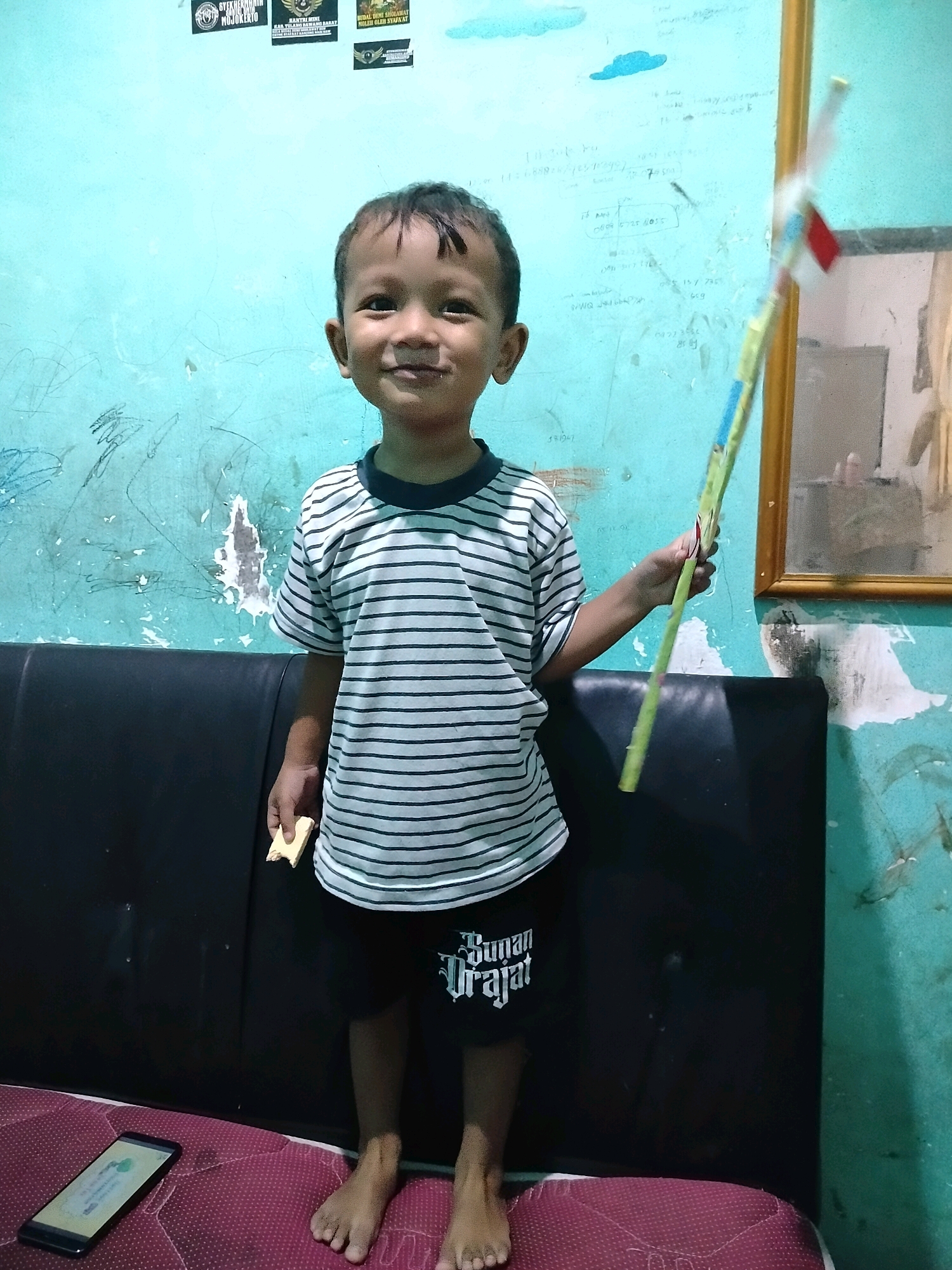 A young boy, appearing to be around five years old, stands on a red-patterned mattress in front of a black sofa. He is wearing a horizontally striped t-shirt and dark shorts with white lettering. In his right hand, he holds a long, green stick with what appears to be a small red and white flag at the top. He is also holding a small piece of food in his left hand. A smartphone is visible on the mattress to the boy's left. The background features a turquoise-colored wall with markings and peeling paint, as well as a framed picture or mirror on the right side. The setting is likely indoors in a residential area in Beloh, Indonesia.