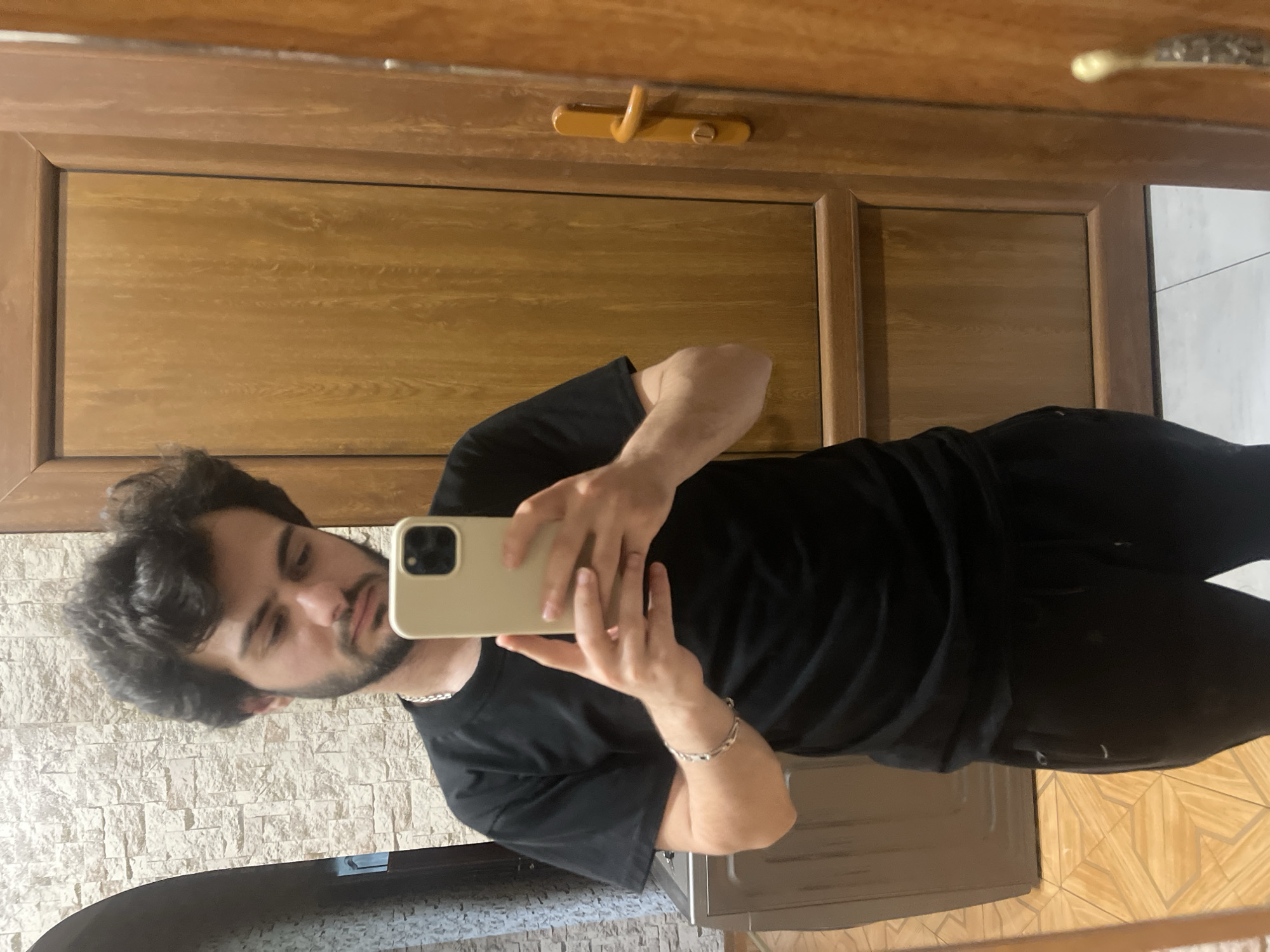 A young man with dark, curly hair and a beard is taking a selfie in a room. He is wearing a black t-shirt and black sweatpants, with a silver chain around his neck and a silver bracelet on his left wrist. He is holding a light-colored smartphone in his right hand to take the picture. The background shows a textured wall to the left, a wooden door with a yellow handle to the right, and a tiled floor. The scene appears to be indoors, likely in a residential setting. There is no visual information to suggest this location is specifically in Tbilisi, Georgia.