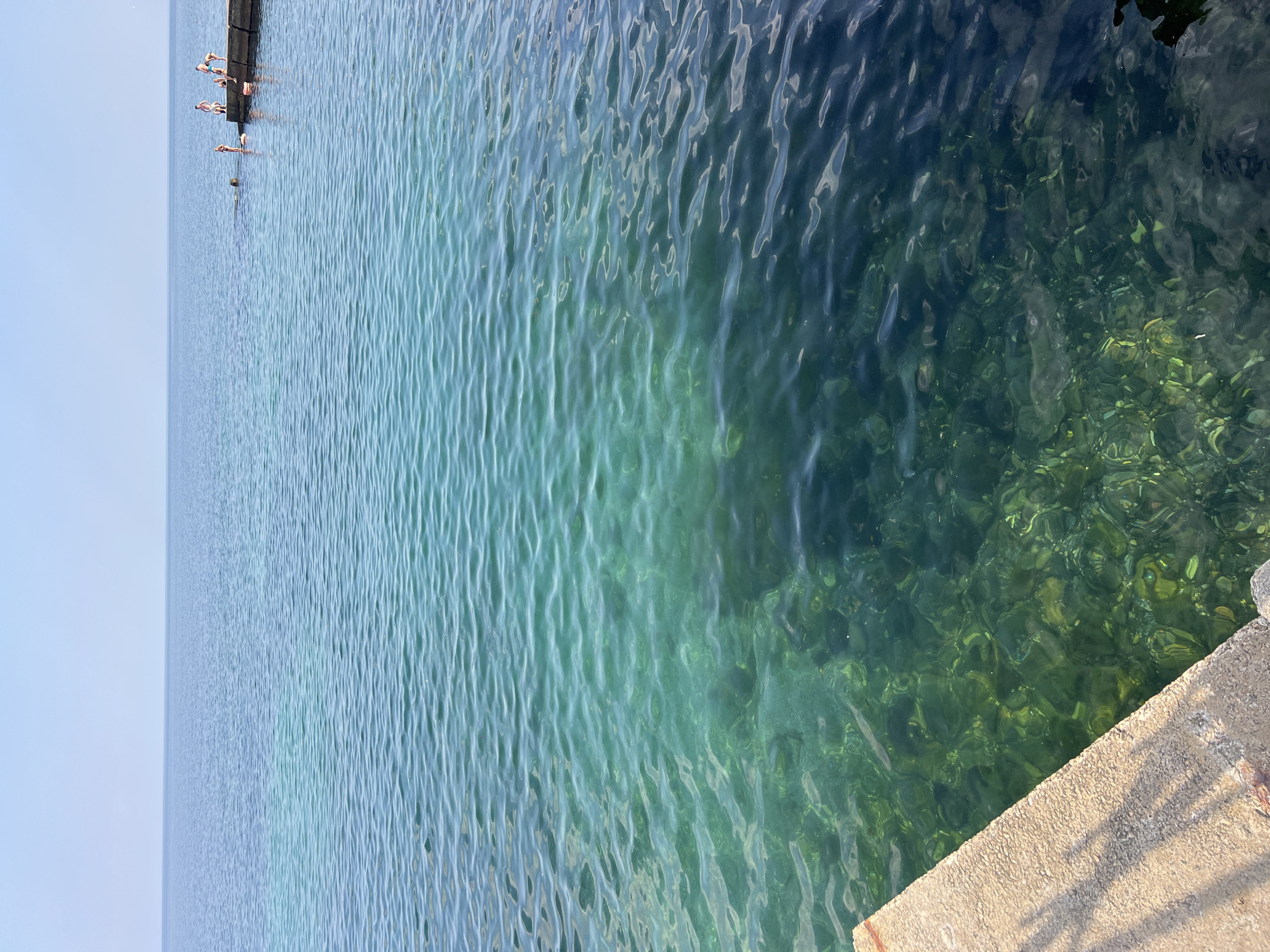 The image shows a view of the Black Sea from a concrete embankment on a clear day. The water transitions from a bright turquoise near the shore to a deep blue further out. The seabed is visible in the shallow, clear water, with rocks and algae. In the distance, a group of people are standing on a concrete pier, some appearing to be in the water or preparing to jump in. The sky is a pale blue with no clouds. The lighting suggests it is daytime, likely late morning or early afternoon. The scene appears to be a public swimming or recreational area in Odesa, Ukraine.