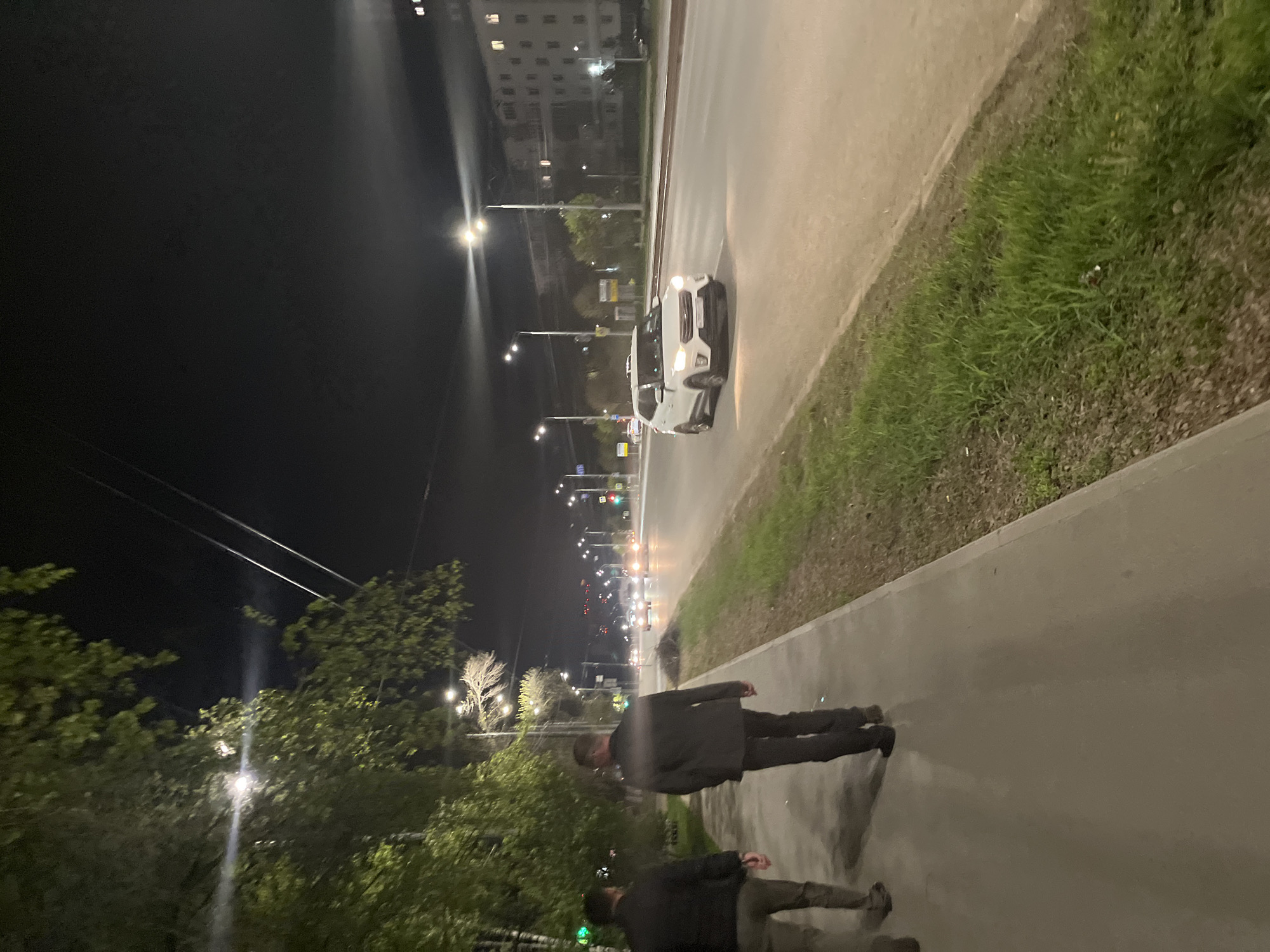 At night, a view of the street in Izhevsk, Russia, shows two people walking on a sidewalk, their reflections visible in the wet pavement. To their right is a road with a white car. Streetlights illuminate the scene, and there are trees and grass alongside the sidewalk. Buildings are visible in the distance along the road.